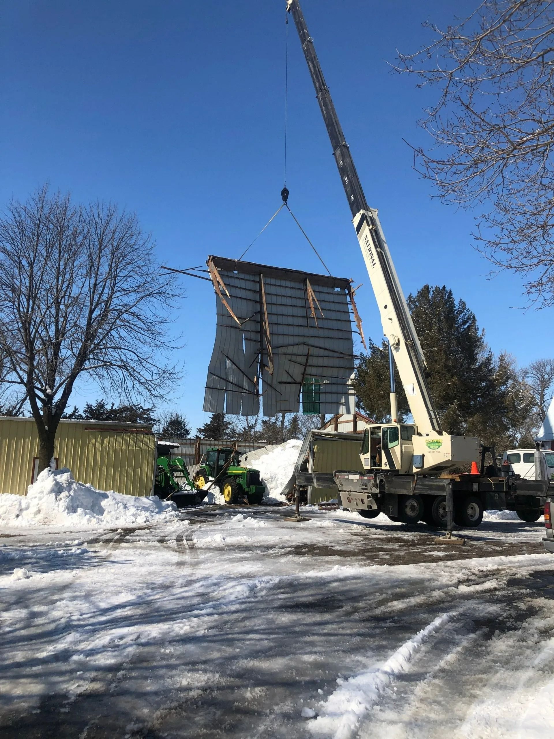 A crane lifting a large, light-colored panel at a construction site covered in snow under a blue sky.