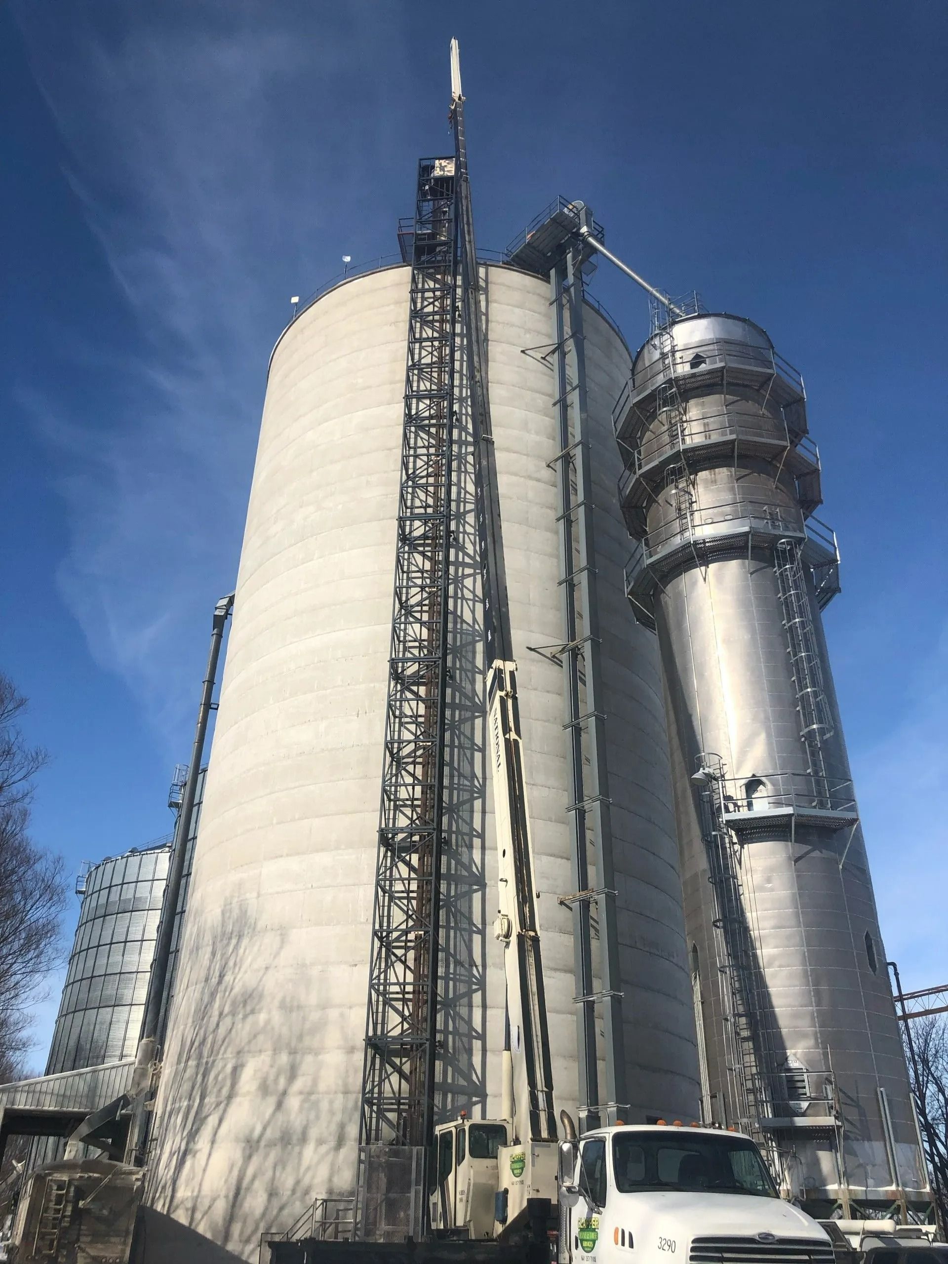 Tall industrial silos and a metal structure against a blue sky, with a white truck in the foreground.