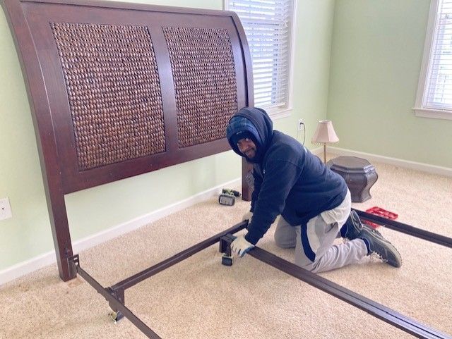A man is kneeling down in front of a bed frame in a bedroom.
