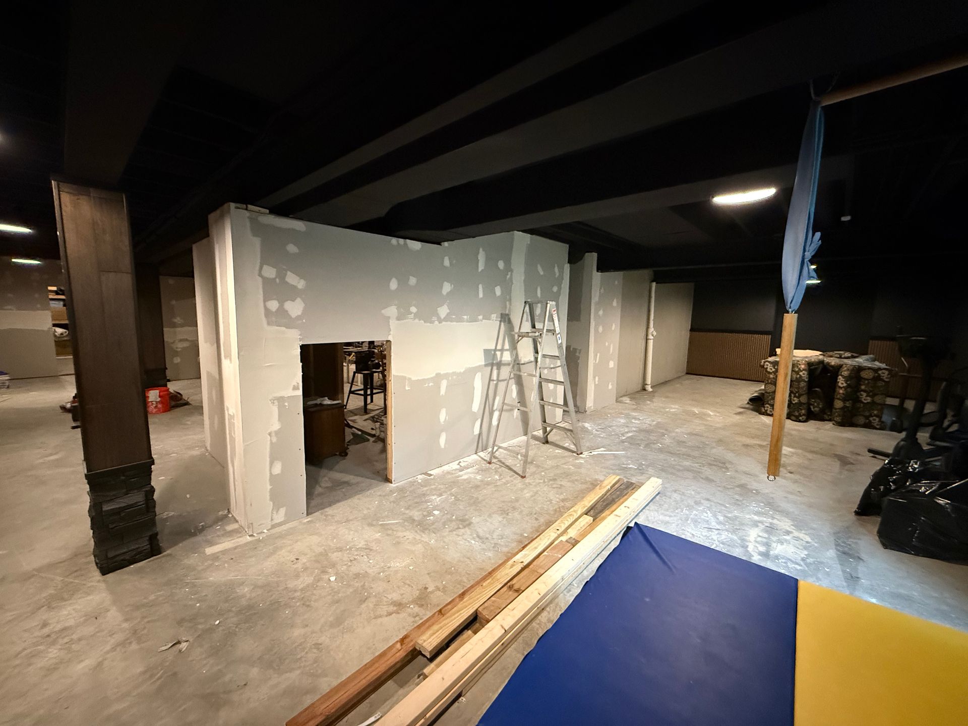 Construction site interior with unfinished drywall, ladder, and lumber under a dark ceiling