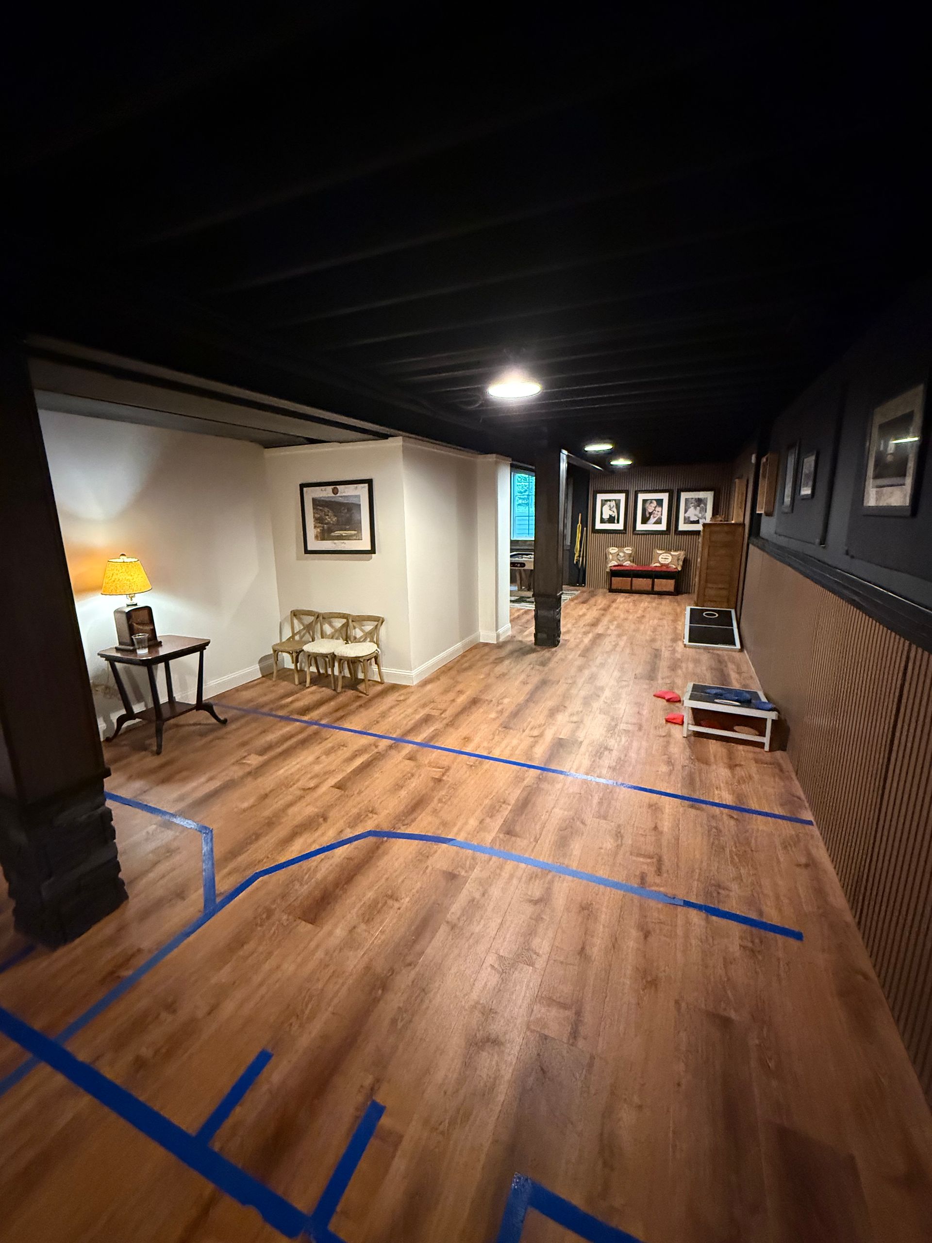 Dim hallway with wood floors, black ceiling, wall art, and blue tape marking the path on the floor.