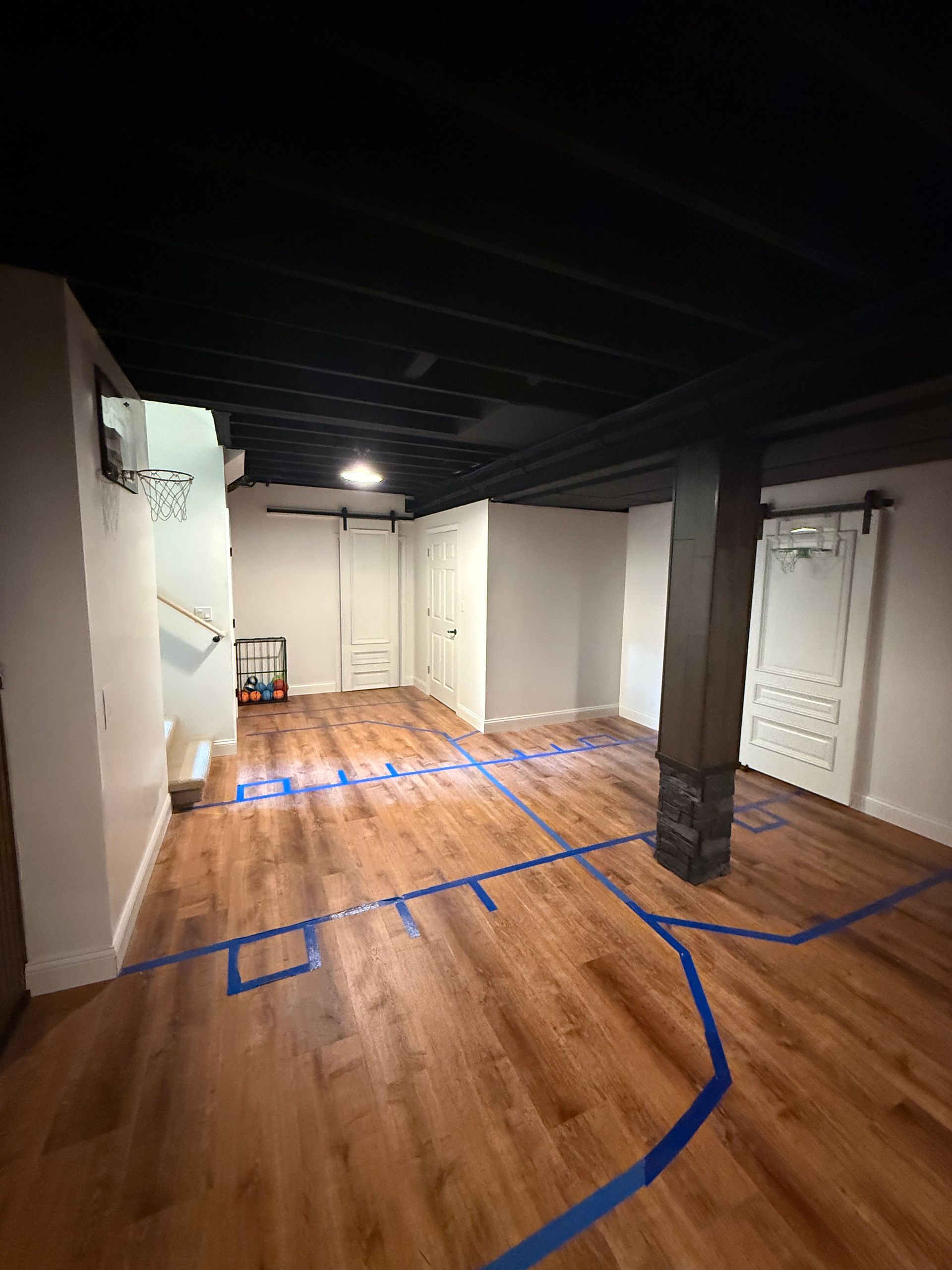 Empty unfinished room with hardwood floors, white walls, and blue tape markings under exposed dark beams