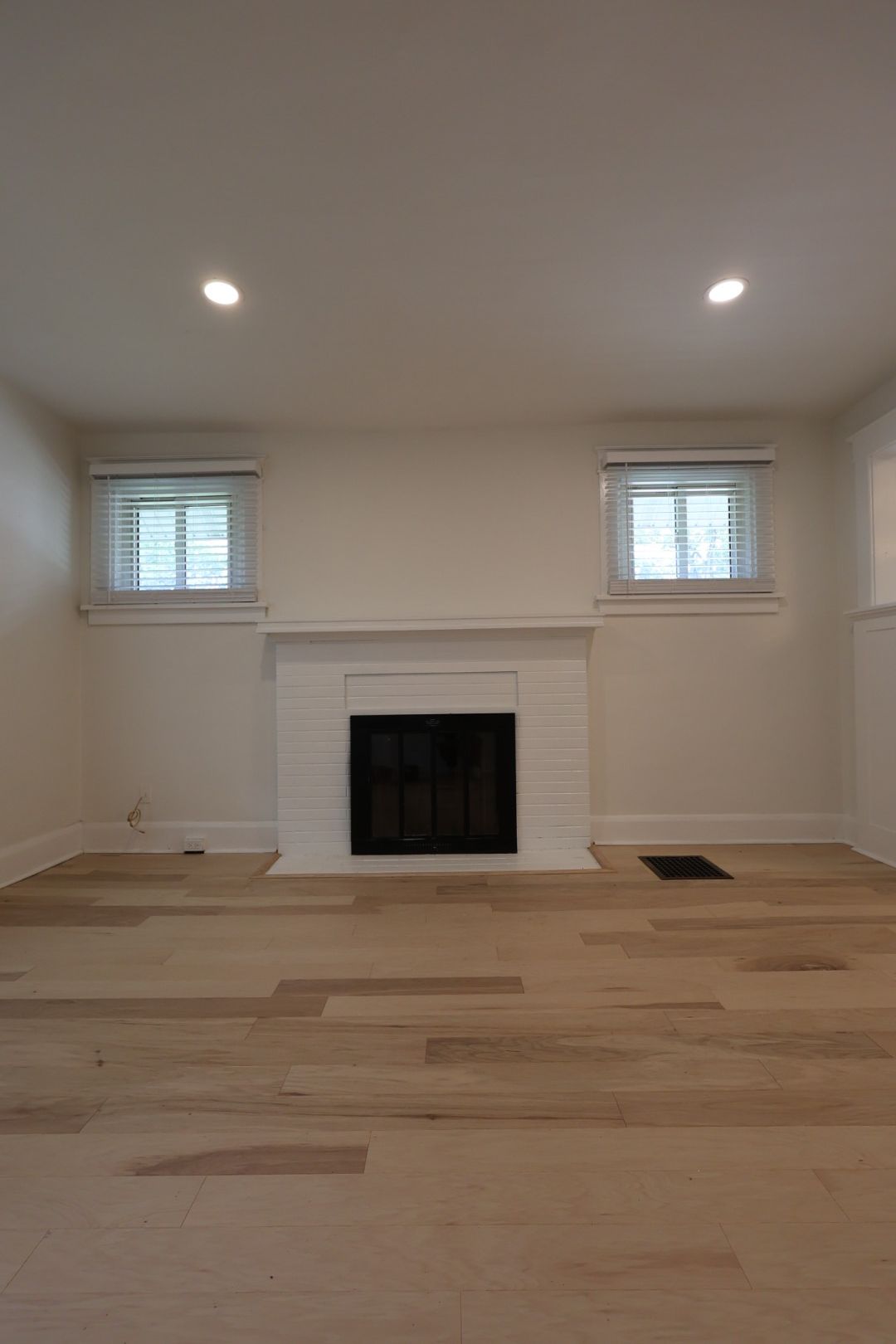 Empty living room with hardwood floors, white walls, small windows, and a fireplace.