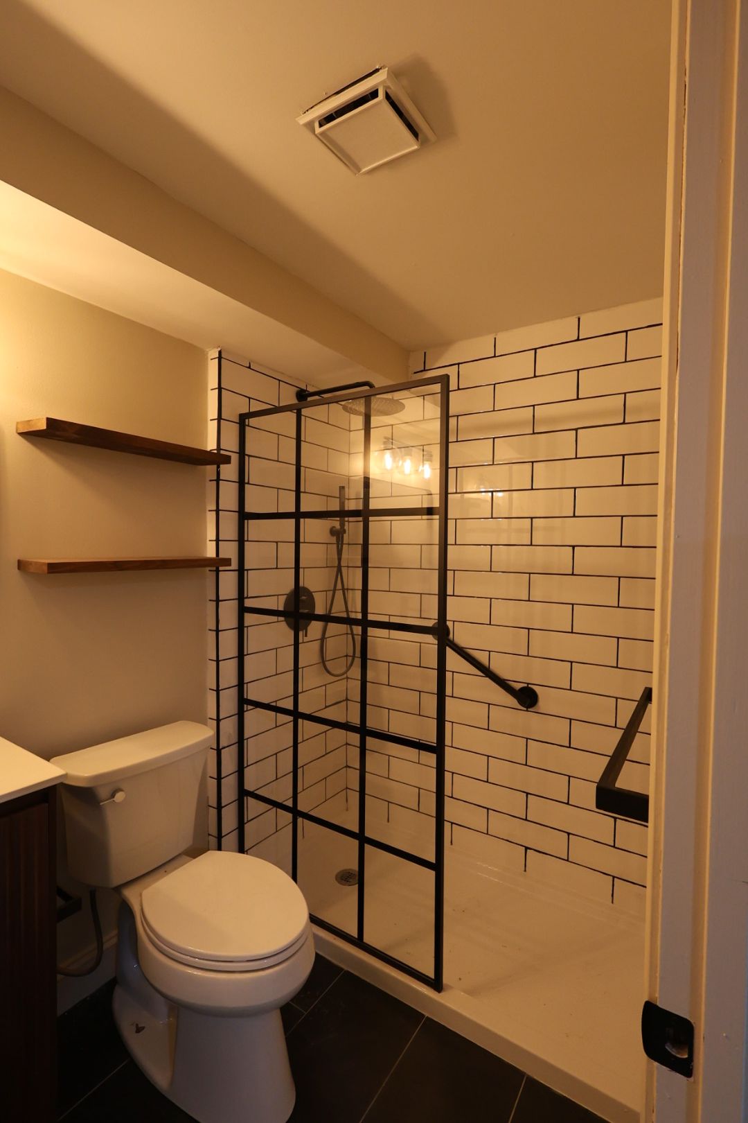 Small bathroom with toilet, shower stall, and white subway-tile wall, lit in warm light.