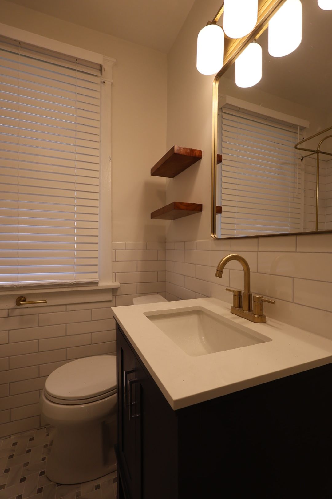 Small bathroom with a white vanity, gold faucet, wall mirror, toilet, and window blinds.