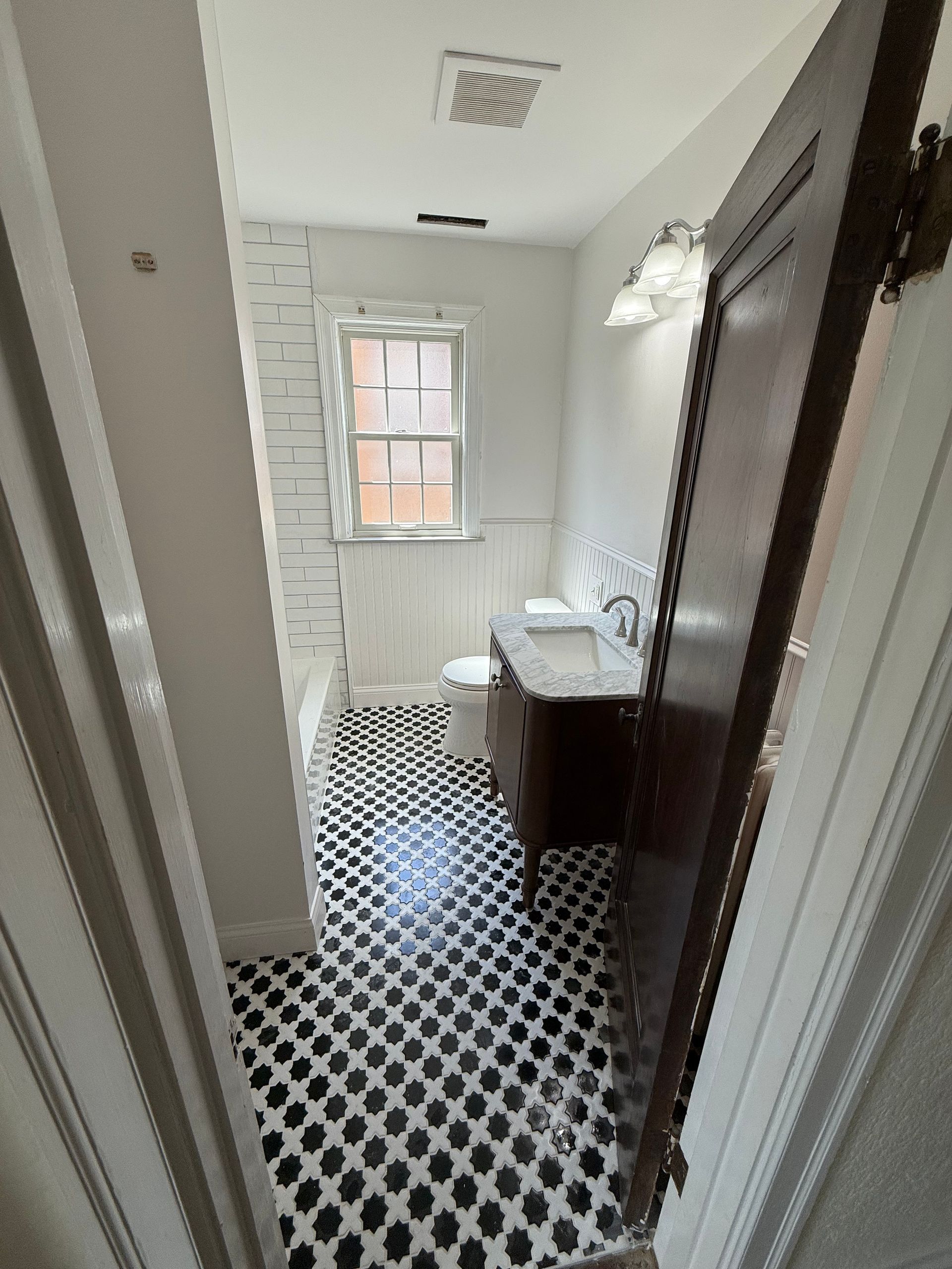 Narrow bathroom with black-and-white tile floor, white walls, window, toilet, and vanity sink.