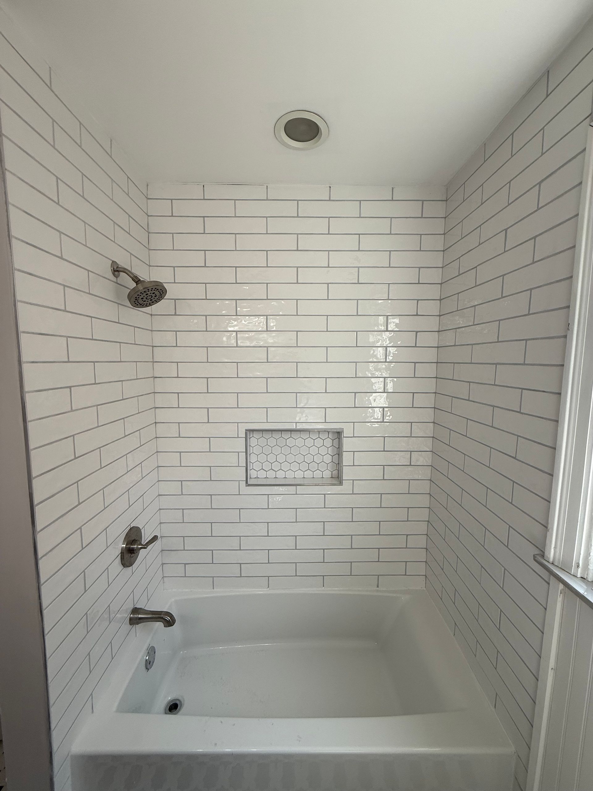 White tiled bathtub and shower alcove with built-in shelf and window light