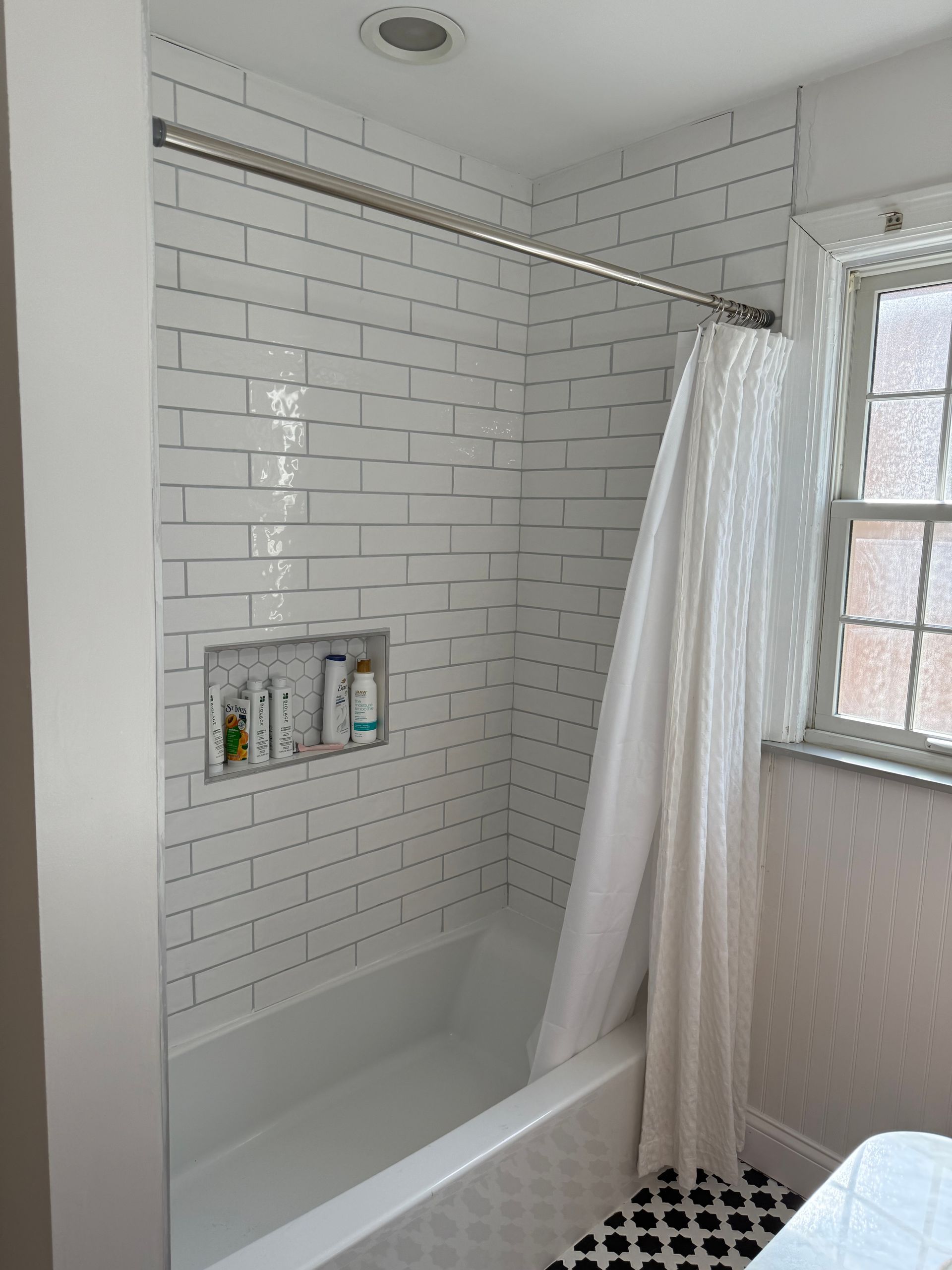 White-tiled bathroom shower with bathtub, built-in shelf, and a sheer curtain by a window