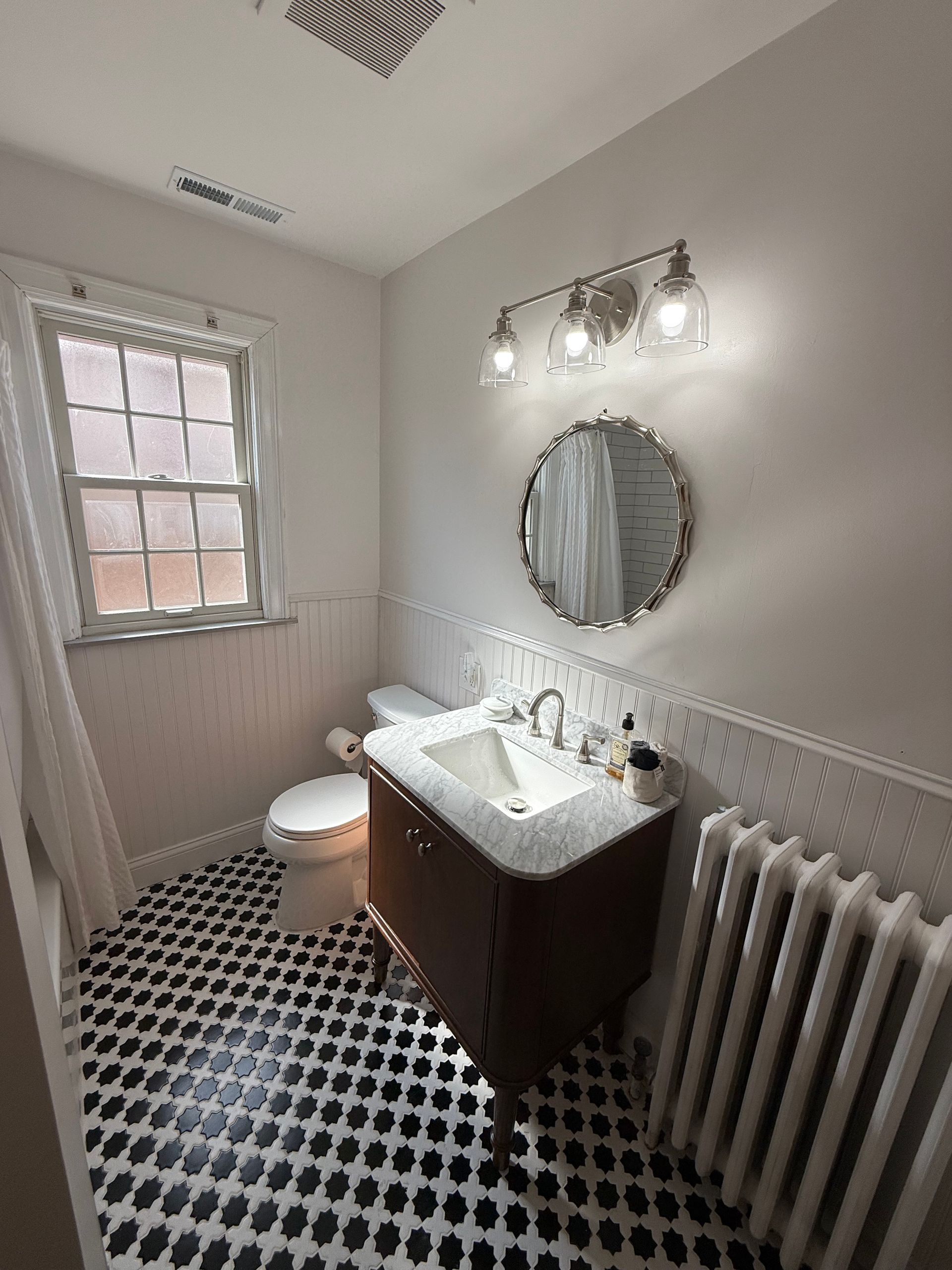 Small bathroom with black-and-white floor tiles, pedestal sink, toilet, window, mirror, and radiator.