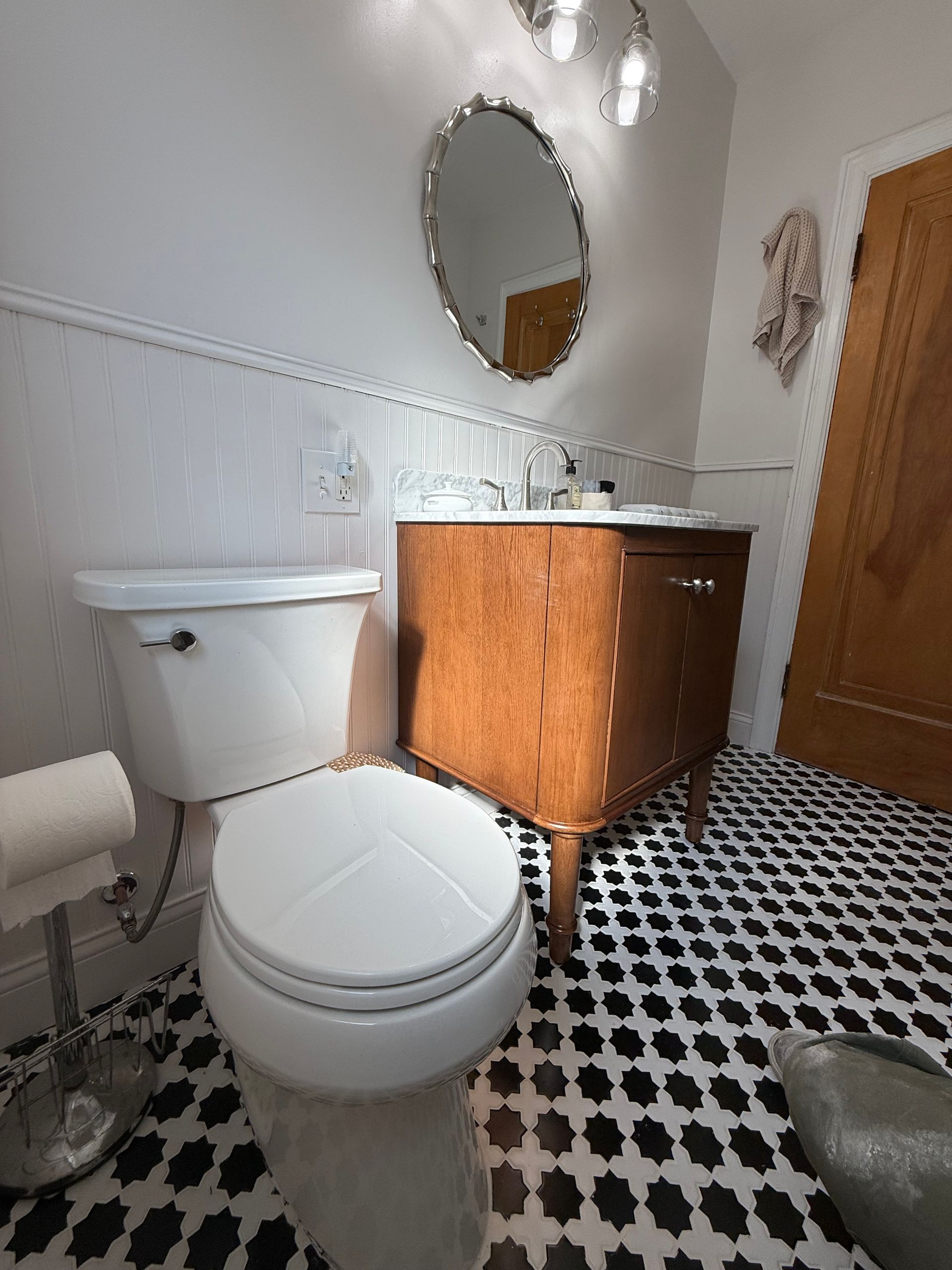 Small bathroom with toilet, wooden vanity, oval mirror, and black-and-white tile floor.