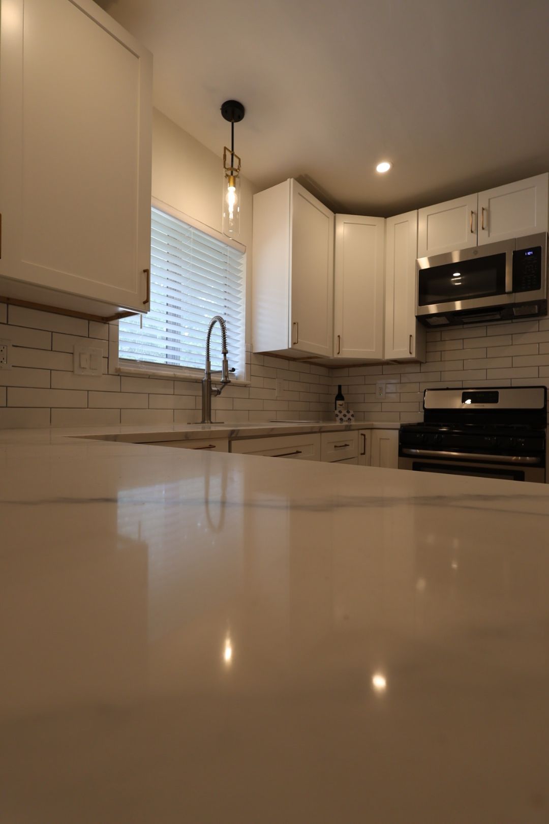 Bright kitchen with white cabinets, marble countertop, window blinds, pendant light, and stainless steel stove.