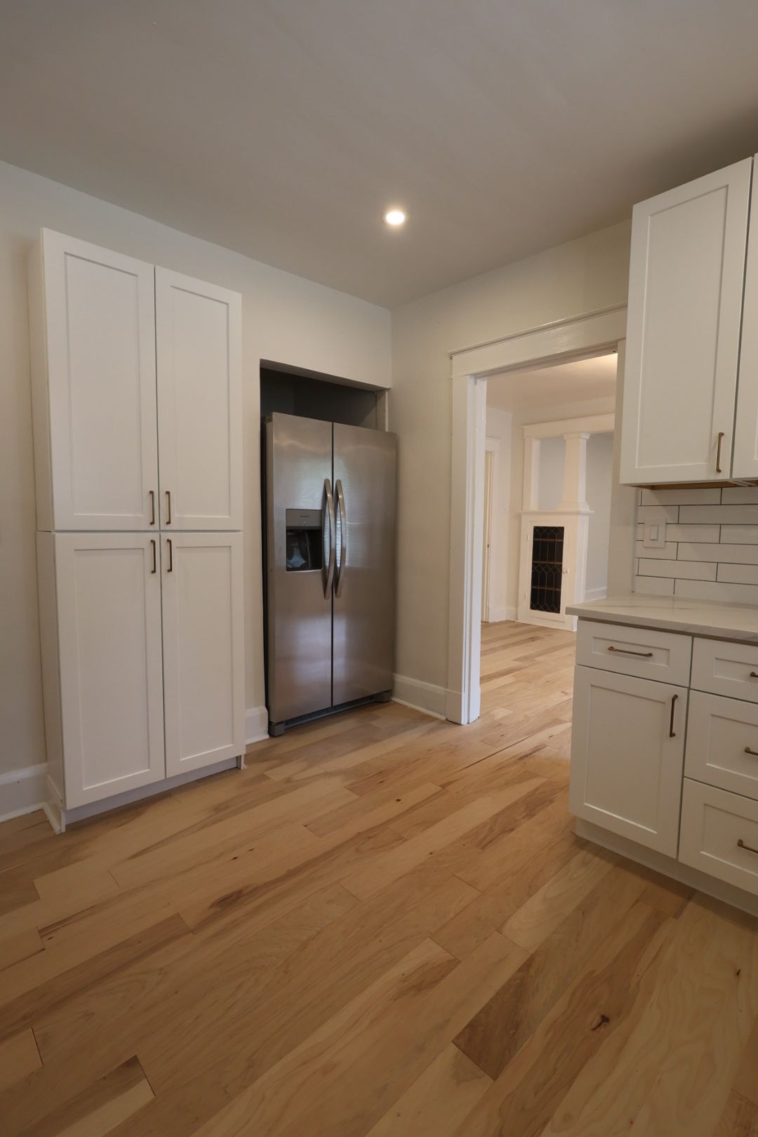 Modern kitchen with white cabinets, stainless steel fridge, and light wood floors leading to a doorway.