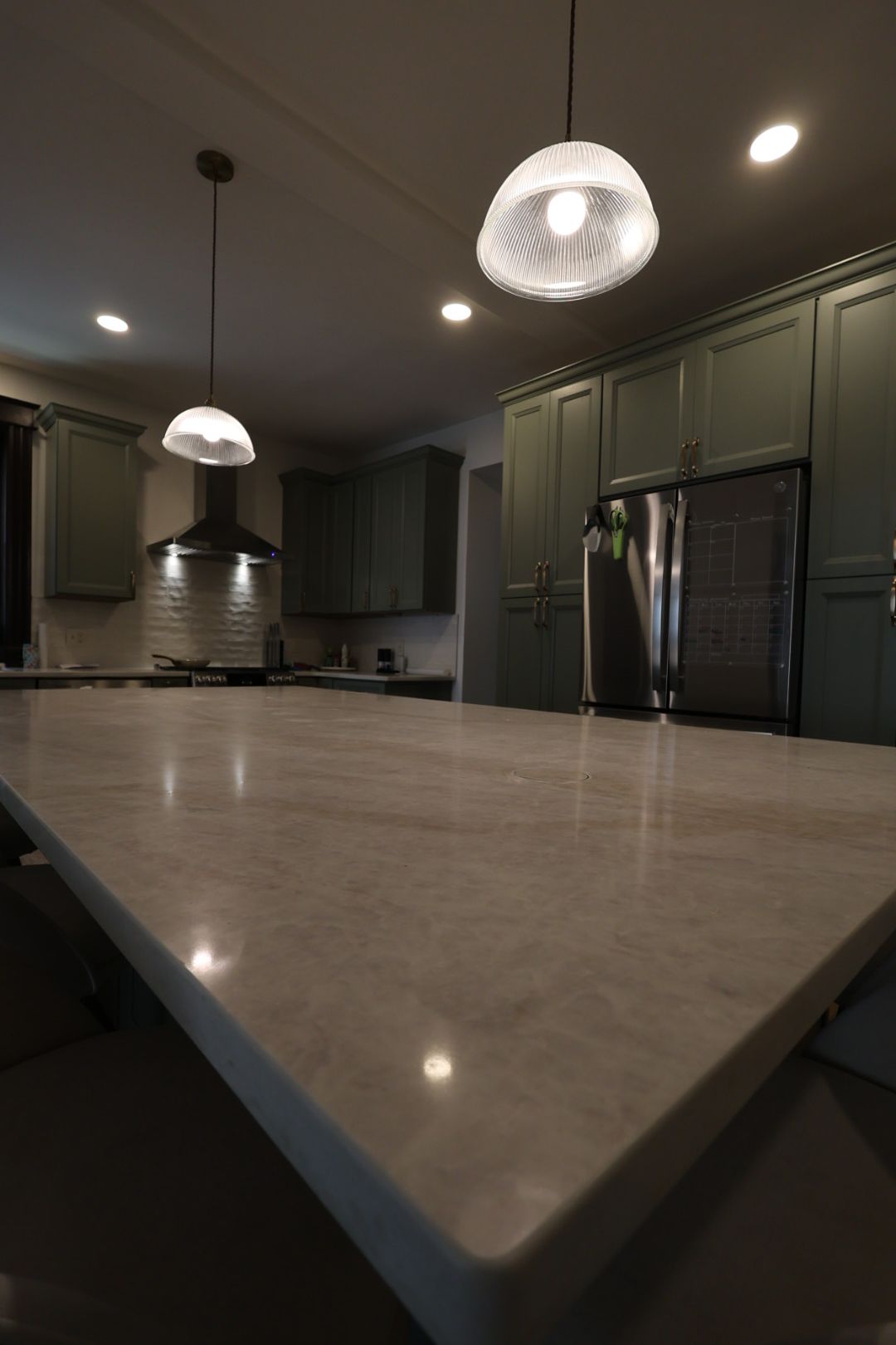 Modern kitchen with a large marble island, dark green cabinets, pendant lights, and a black refrigerator.