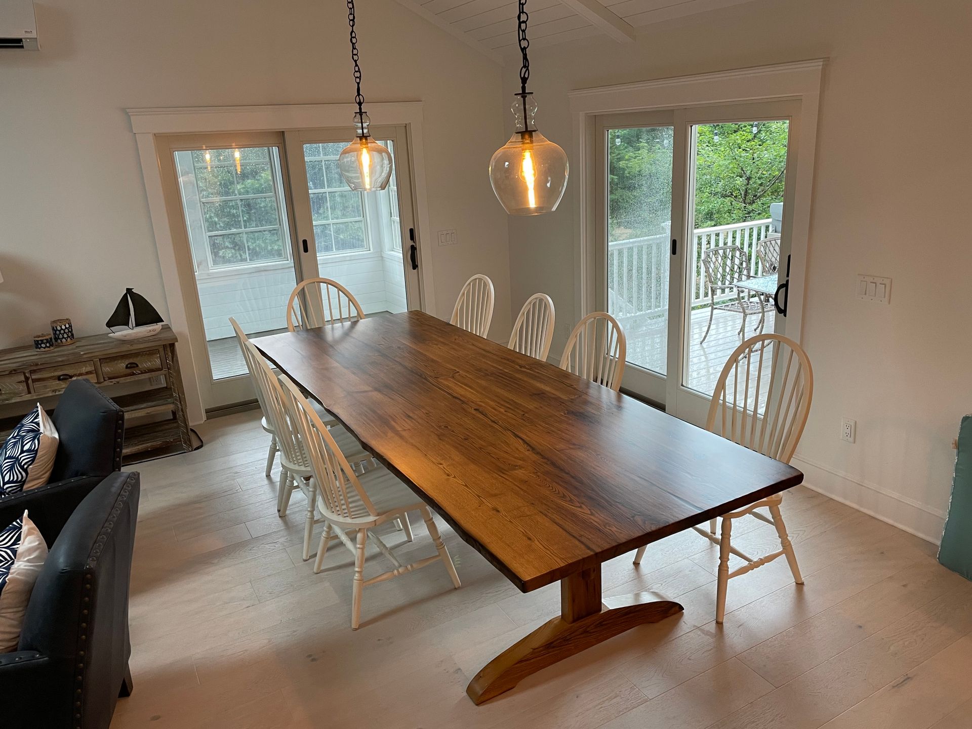 A long wooden table with white chairs in a living room with sliding glass doors.