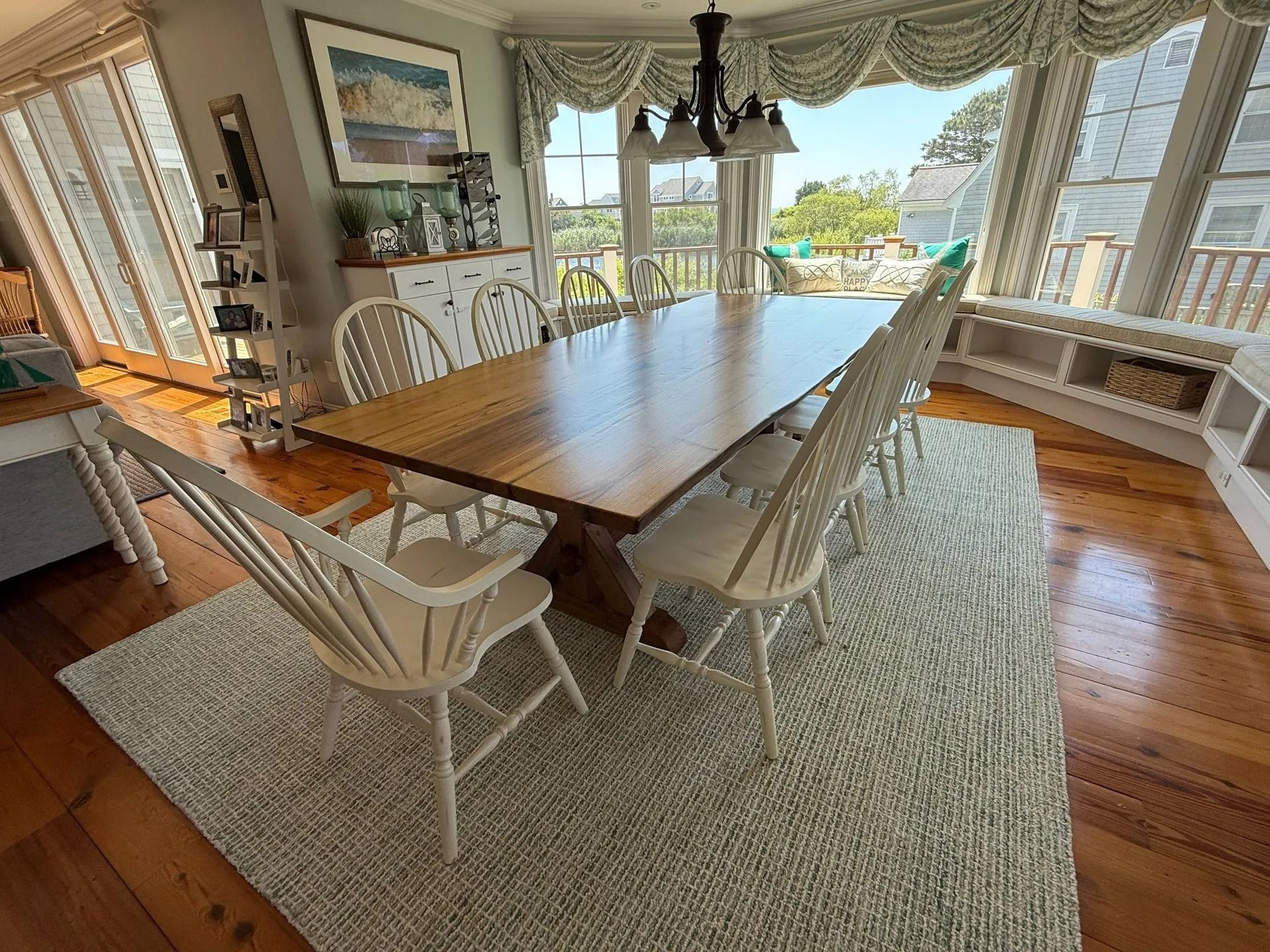 Bright dining room with a long wooden table, white chairs, and large windows overlooking water and greenery