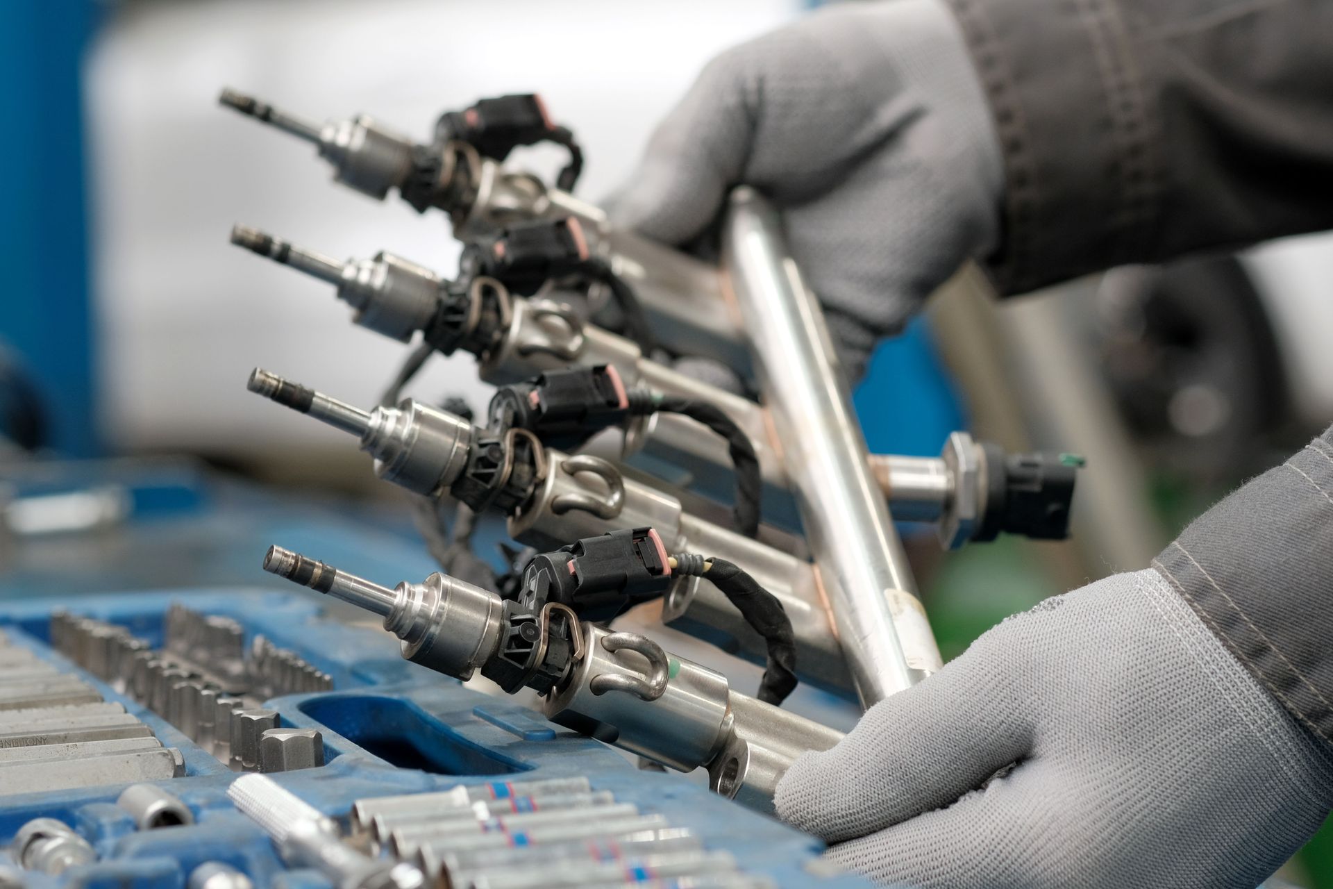 Hands in gloves holding a fuel injector rail assembly, tools in a tray.