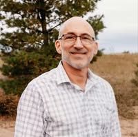 A man wearing glasses and a plaid shirt is standing in front of a tree.