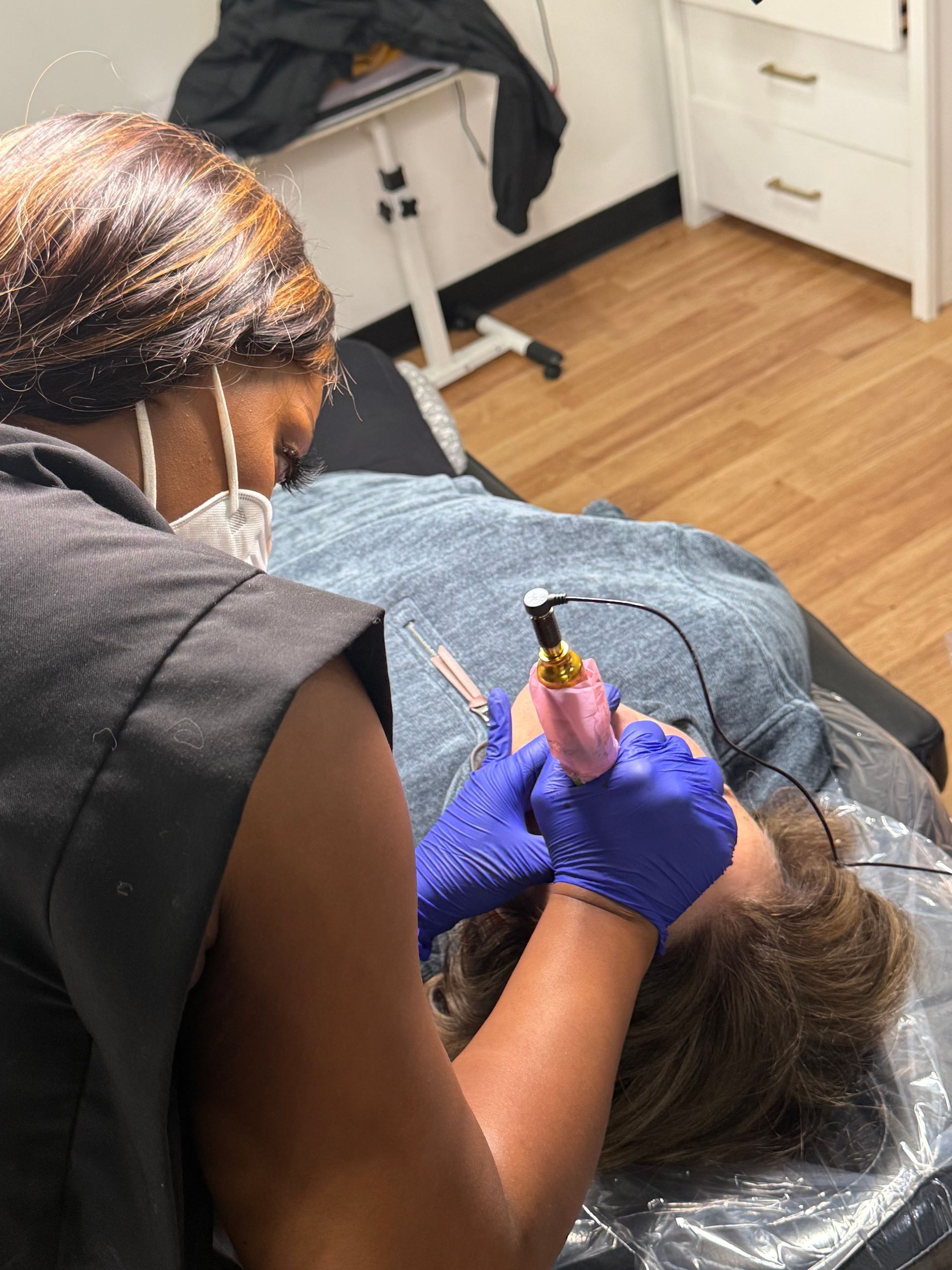 Person performing cosmetic tattooing on a client's face. Both wear gloves. Setting is a treatment room.