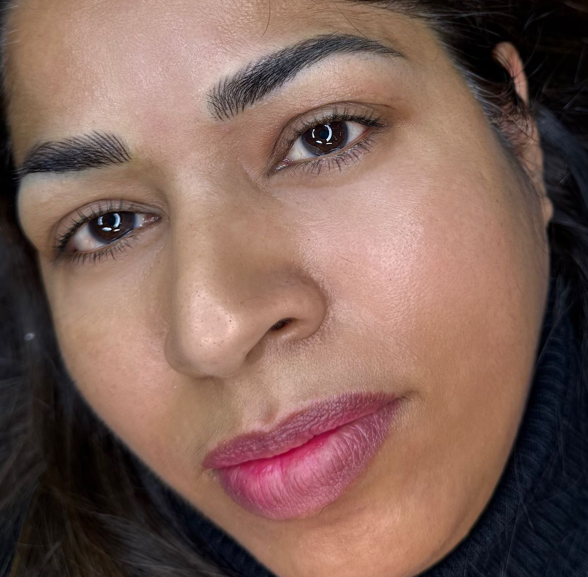 Woman with dark hair, looking at the camera. She has filled-in eyebrows and pink lipstick.