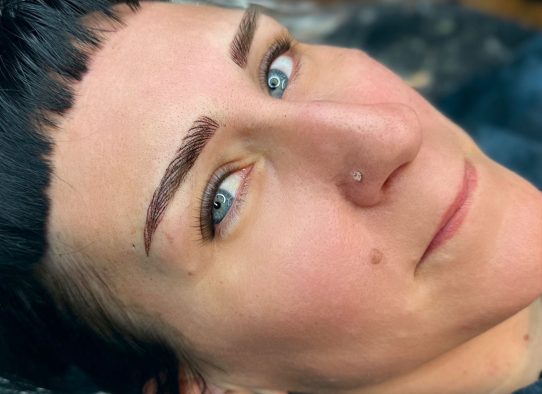 Woman with new eyebrow tattoos, blue eyes, and black hair; lying down.