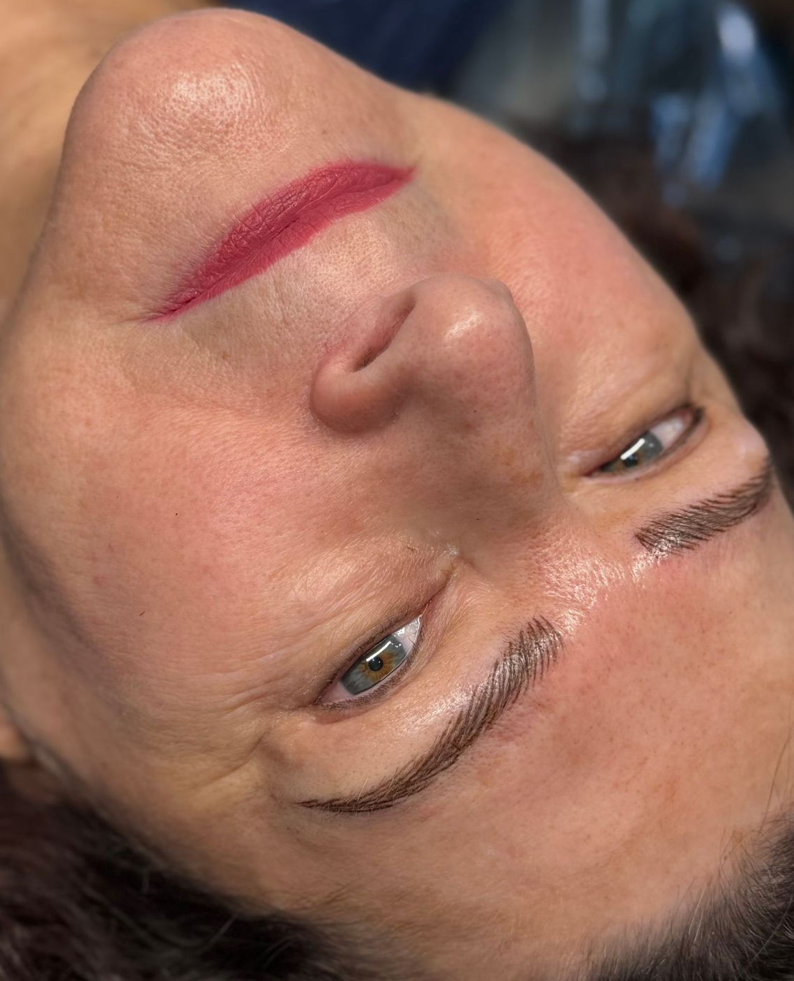 Woman with red lipstick and newly tattooed eyebrows, looking upward.