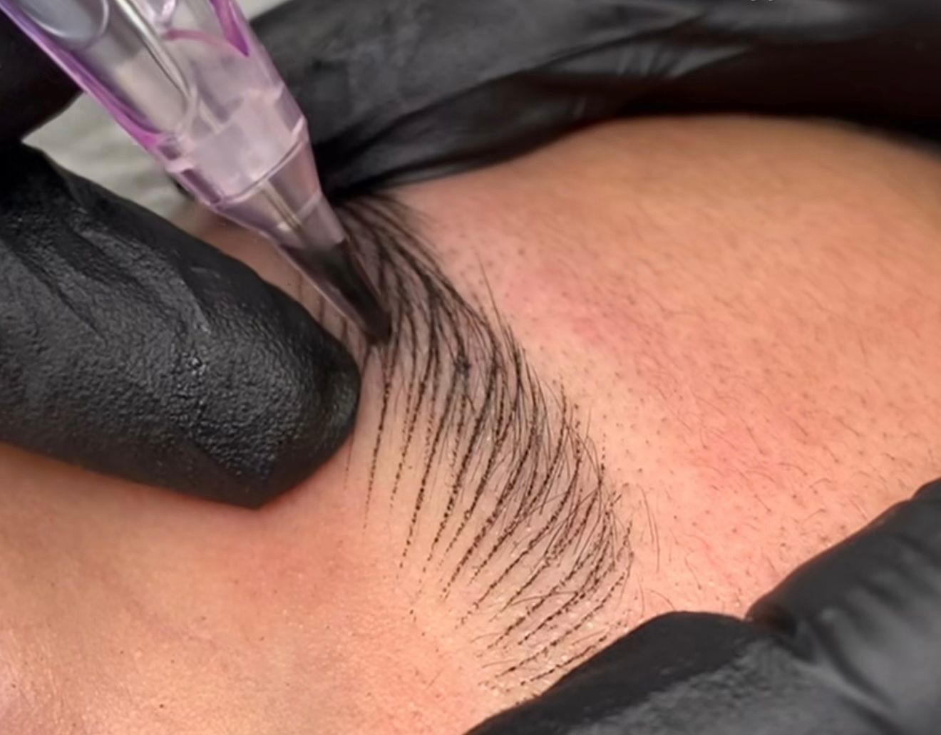 Close-up of eyebrow tattooing: black-gloved hand using a pen-like device to draw thin lines on the brow.
