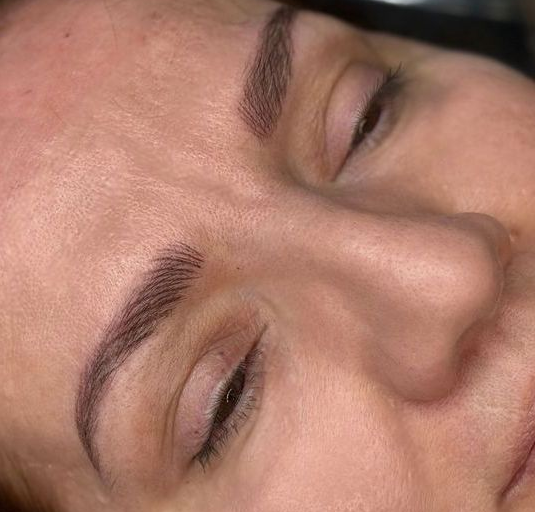 Close-up of a person's face with new eyebrow tattoos; eyes closed.