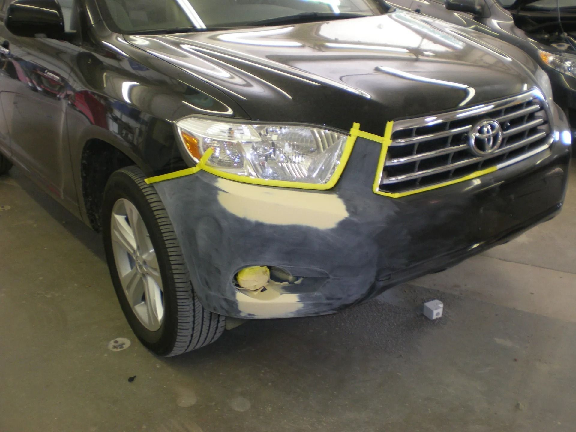 Car's front bumper being repaired; primer is visible. Yellow tape outlines the repair area on a black Toyota Highlander.