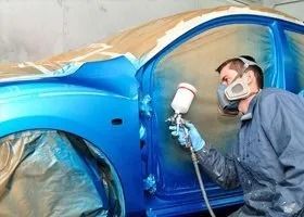 Man in a mask sprays blue paint on a car door in a workshop.
