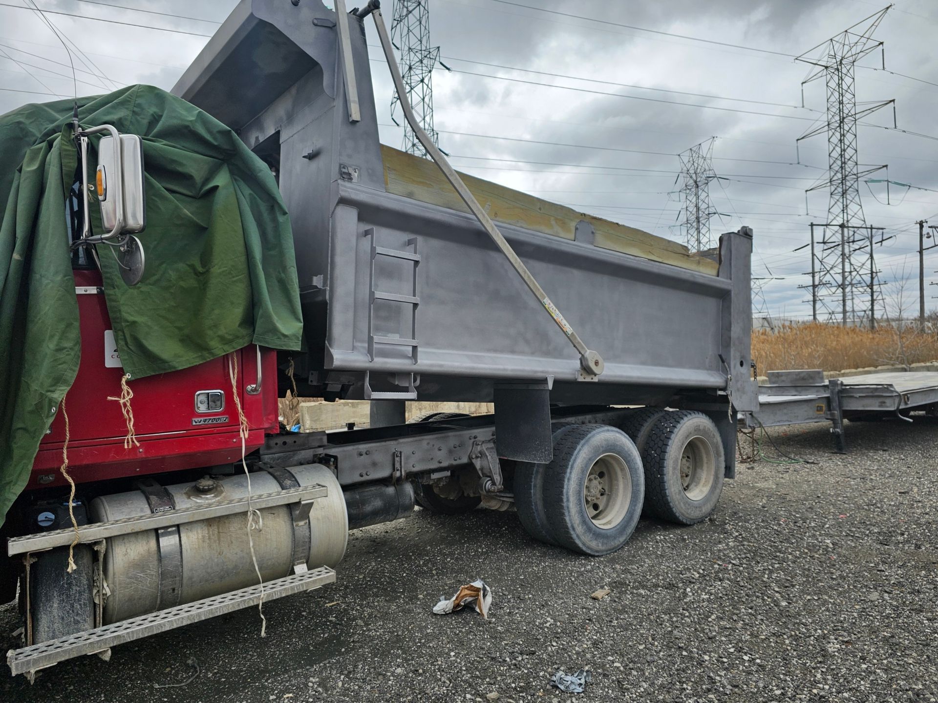 A dump truck with a green tarp on top is parked in a gravel lot.