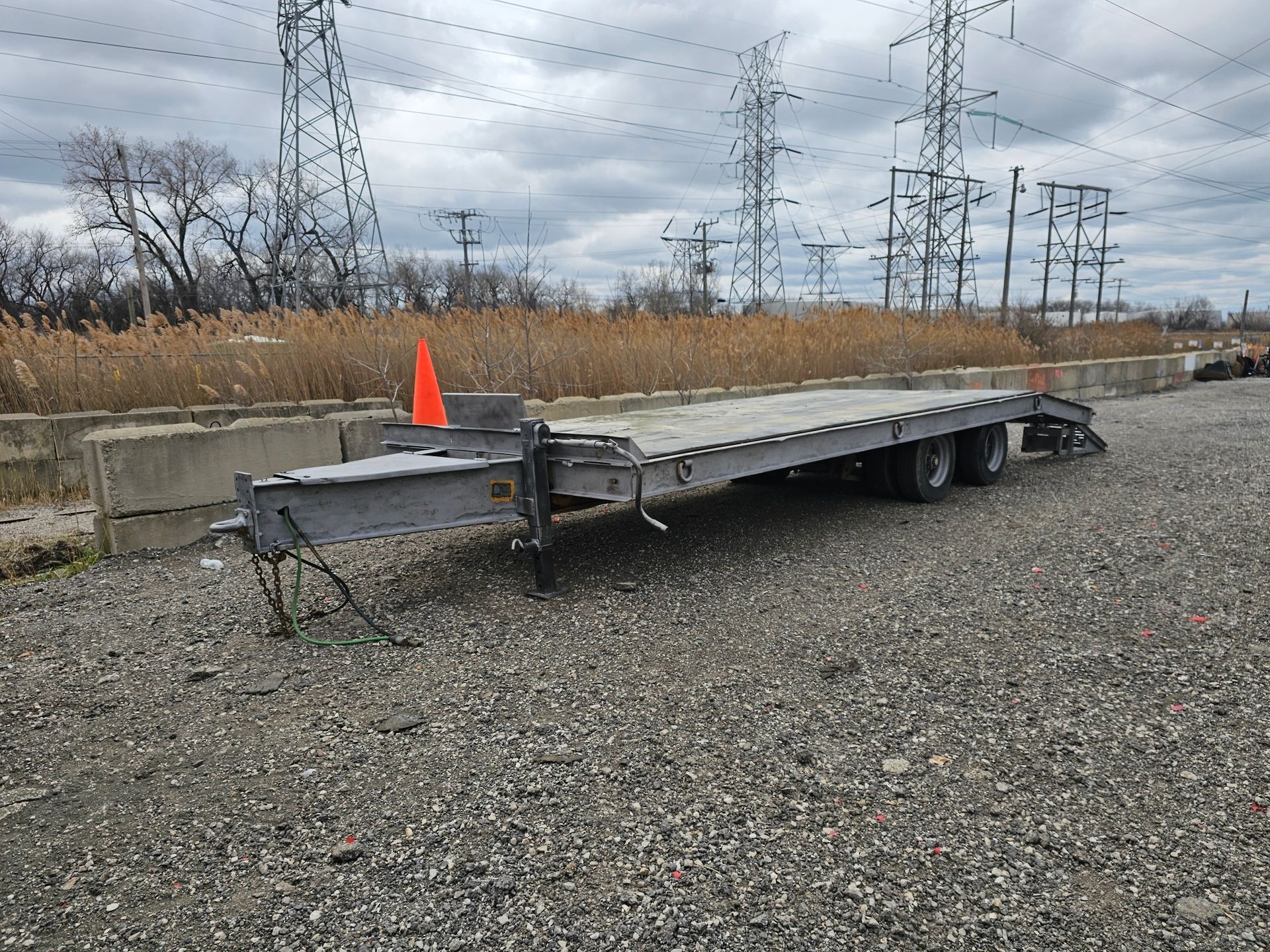 A flatbed trailer with an orange cone attached to it is parked in a gravel lot.