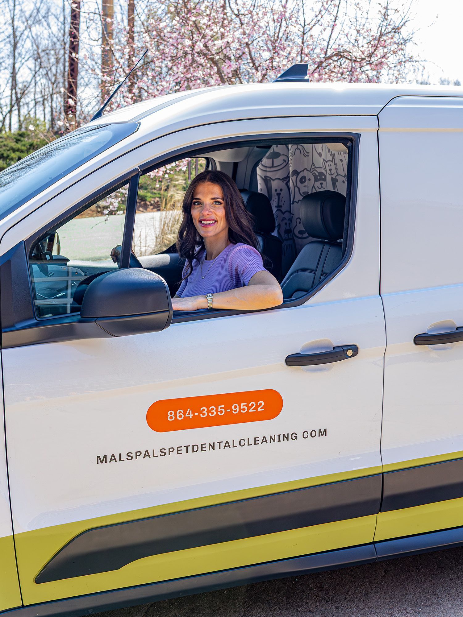 Woman in purple scrubs smiles from a white dental van. The van has business information on the side.