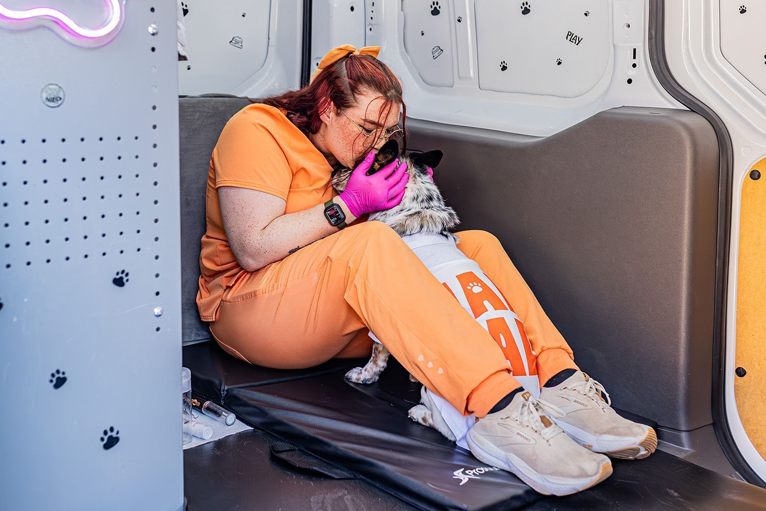 Woman in orange scrubs pets a dog inside a van; the dog wears a white shirt.