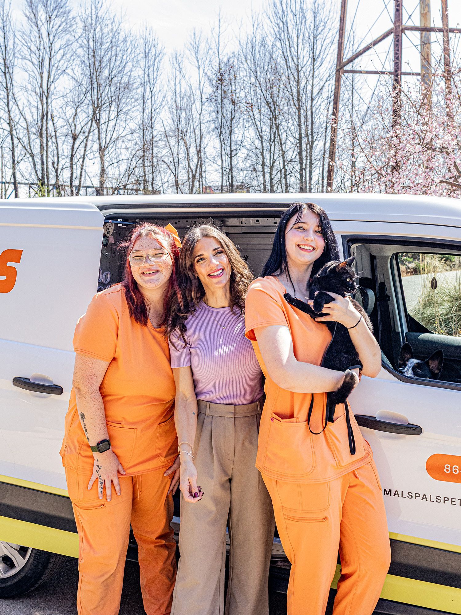 Three people and a small black dog in front of a white van with orange text. Two people wear orange scrubs, one a purple shirt.