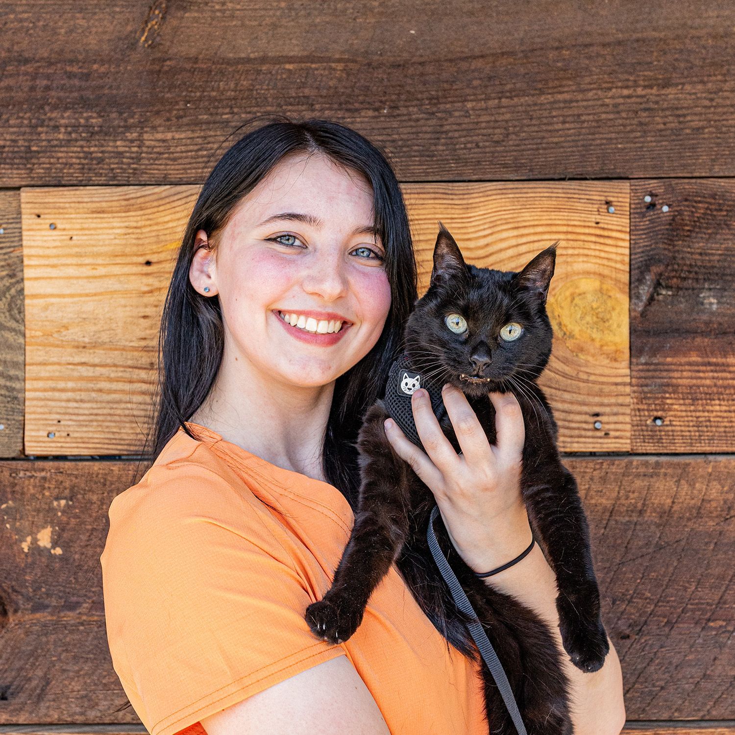 Woman in orange shirt smiles, holding a black cat with leash; wooden wall background.