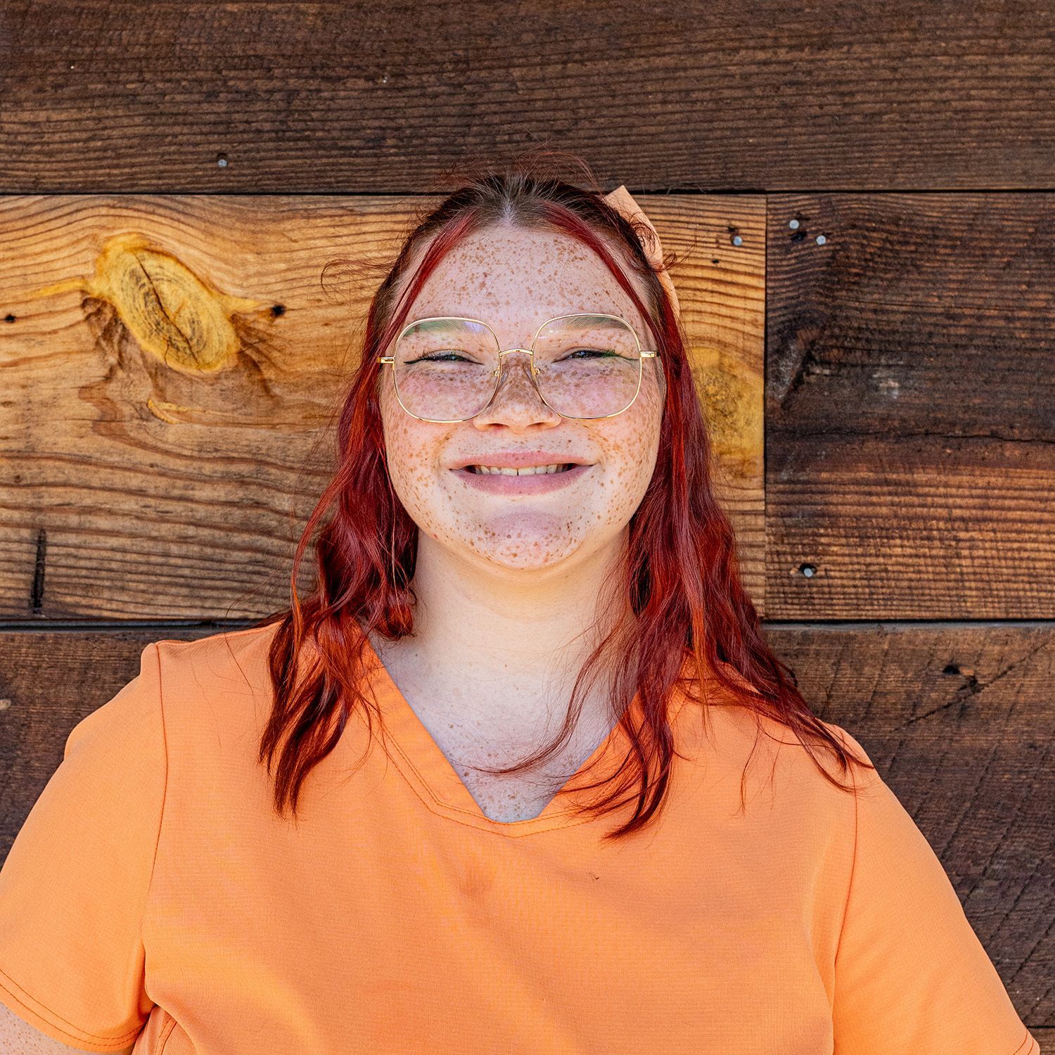 Person with red hair and glasses smiles in front of a wood wall, wearing an orange shirt.