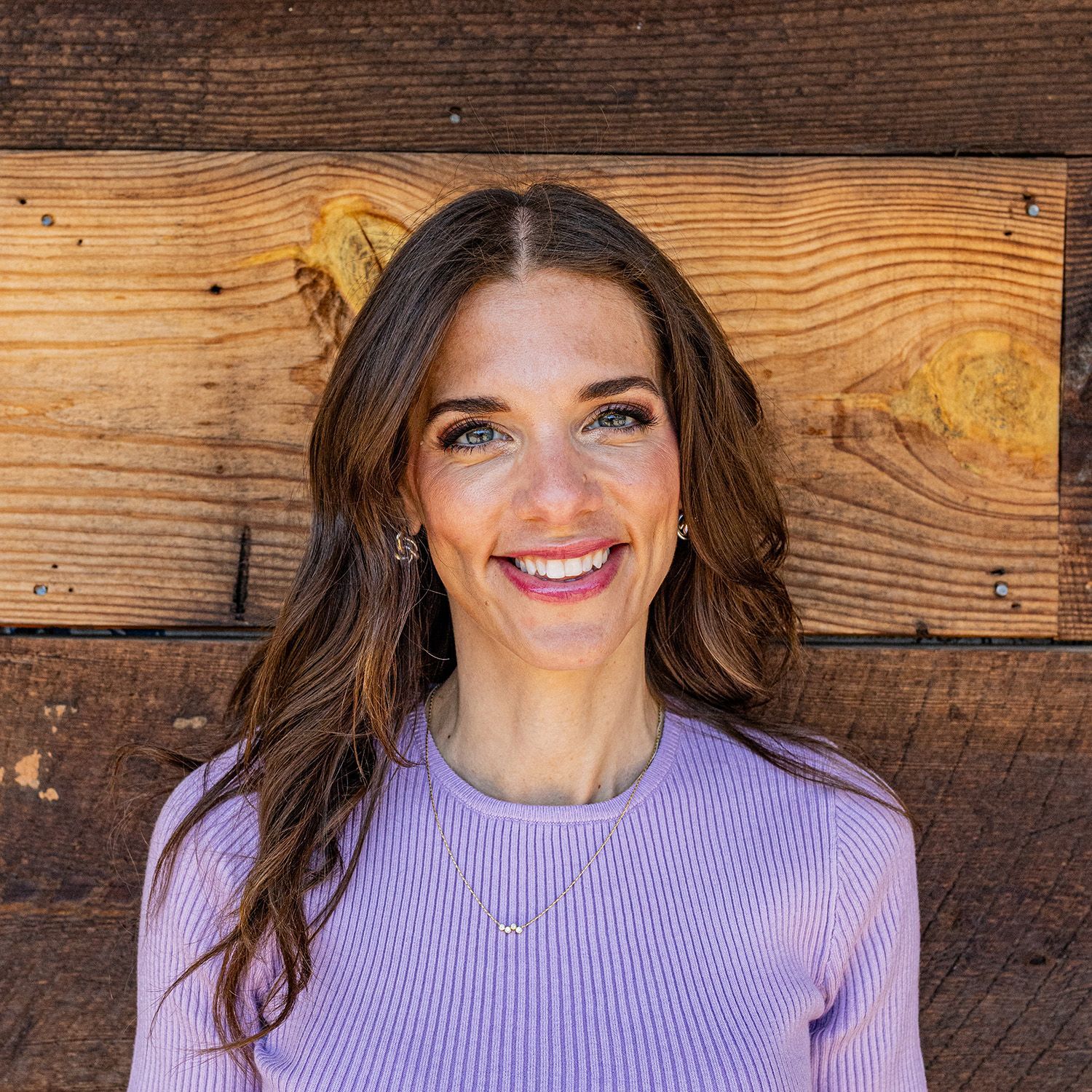 Woman with long brown hair smiles, wearing a lavender sweater, in front of a wood-plank wall.