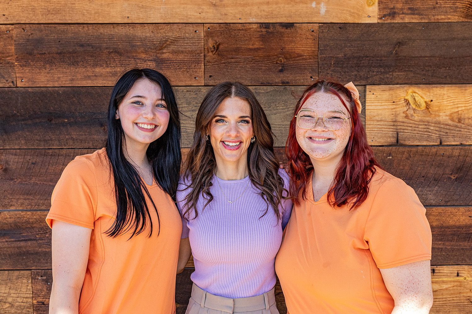 Three women smiling in front of a wood wall; two in orange shirts, one in a purple top and tan pants.