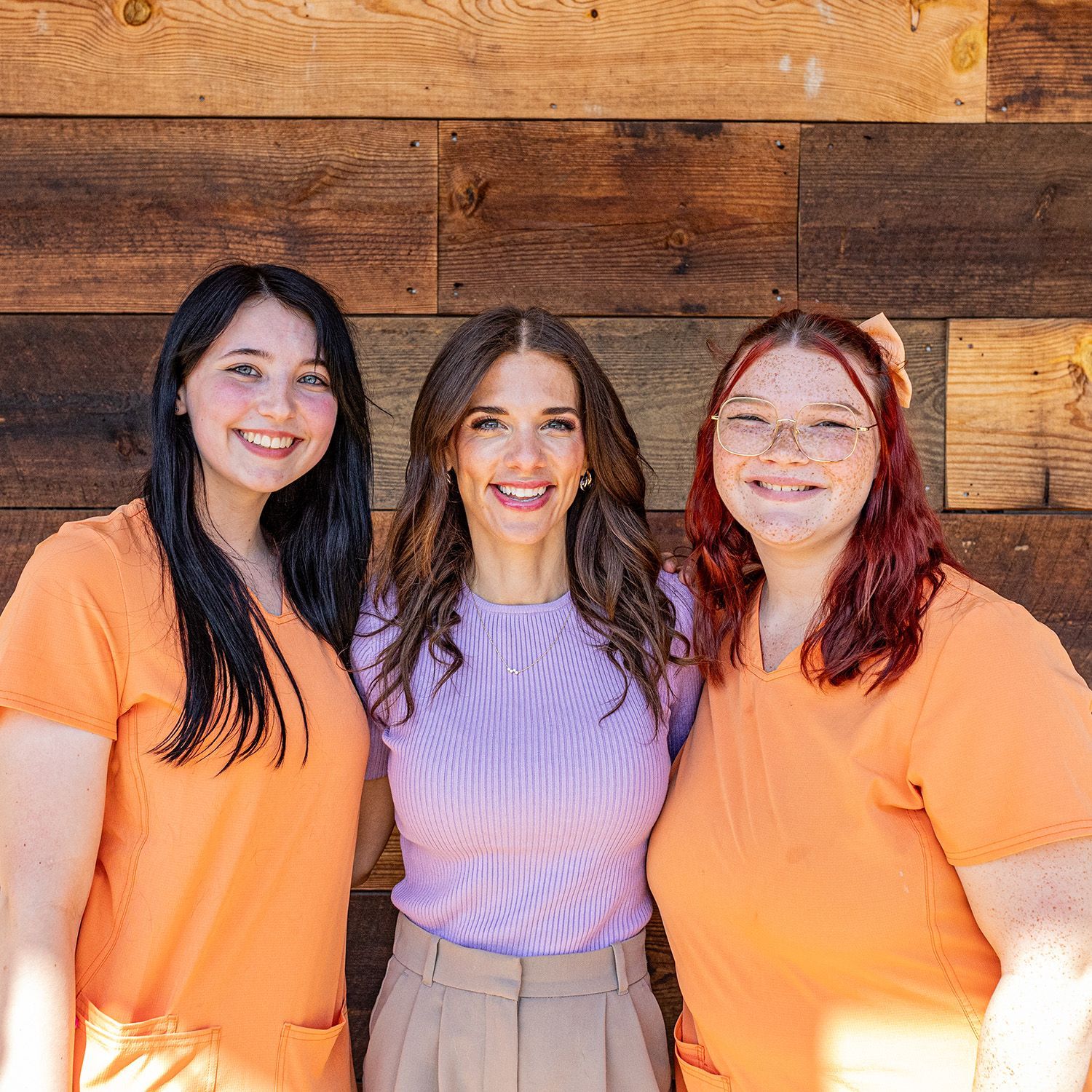 Three women smiling, posing in front of a wood-paneled wall. One wears purple, the others orange.