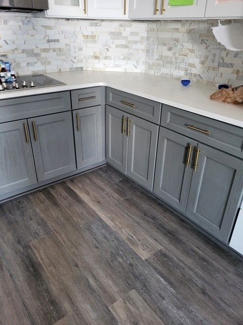 Gray kitchen cabinets with gold handles and wood-look flooring. White countertop with a tile backsplash.