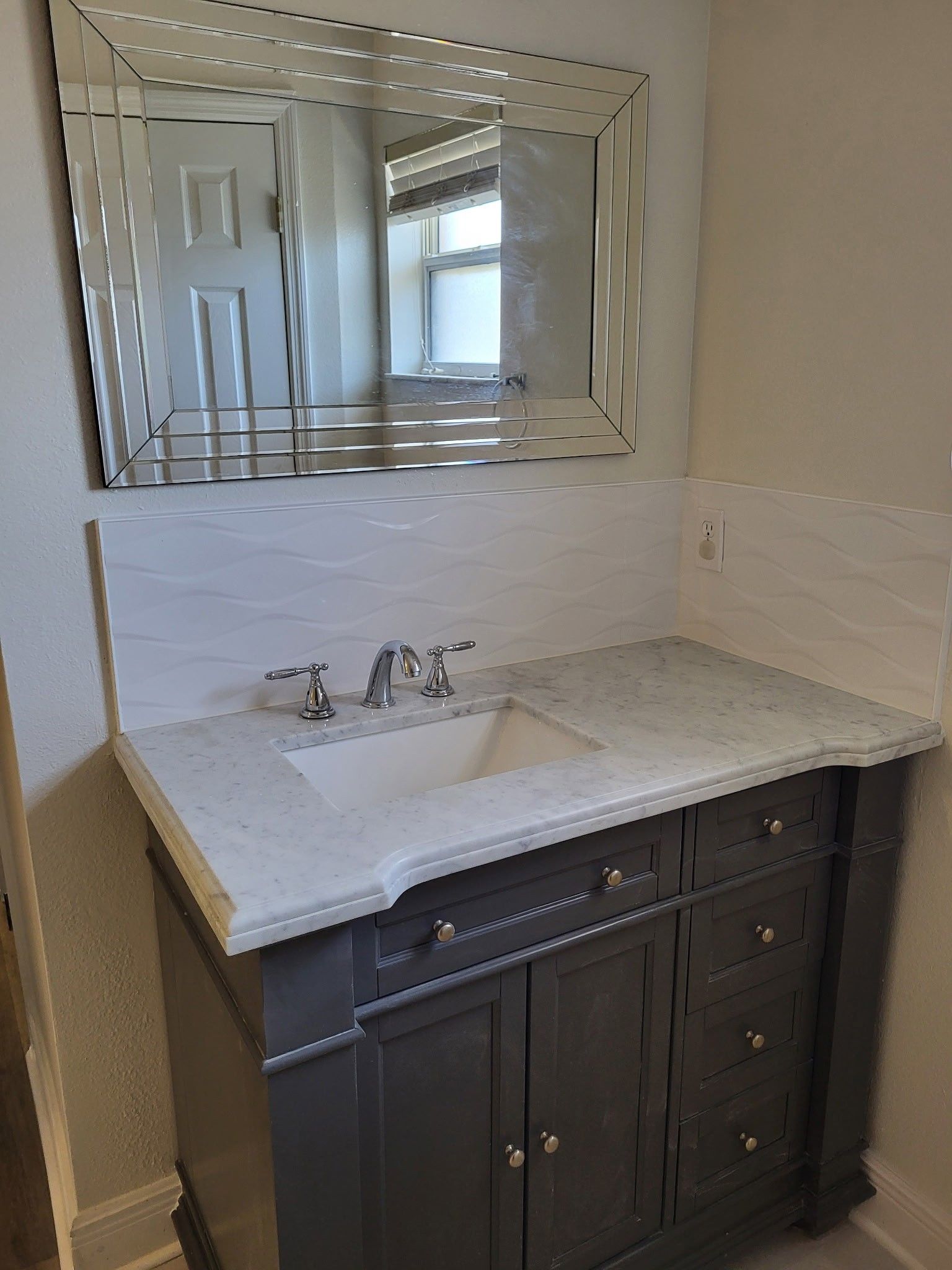 Gray vanity with marble countertop and silver faucet. White tile backsplash, decorative mirror, bright room.