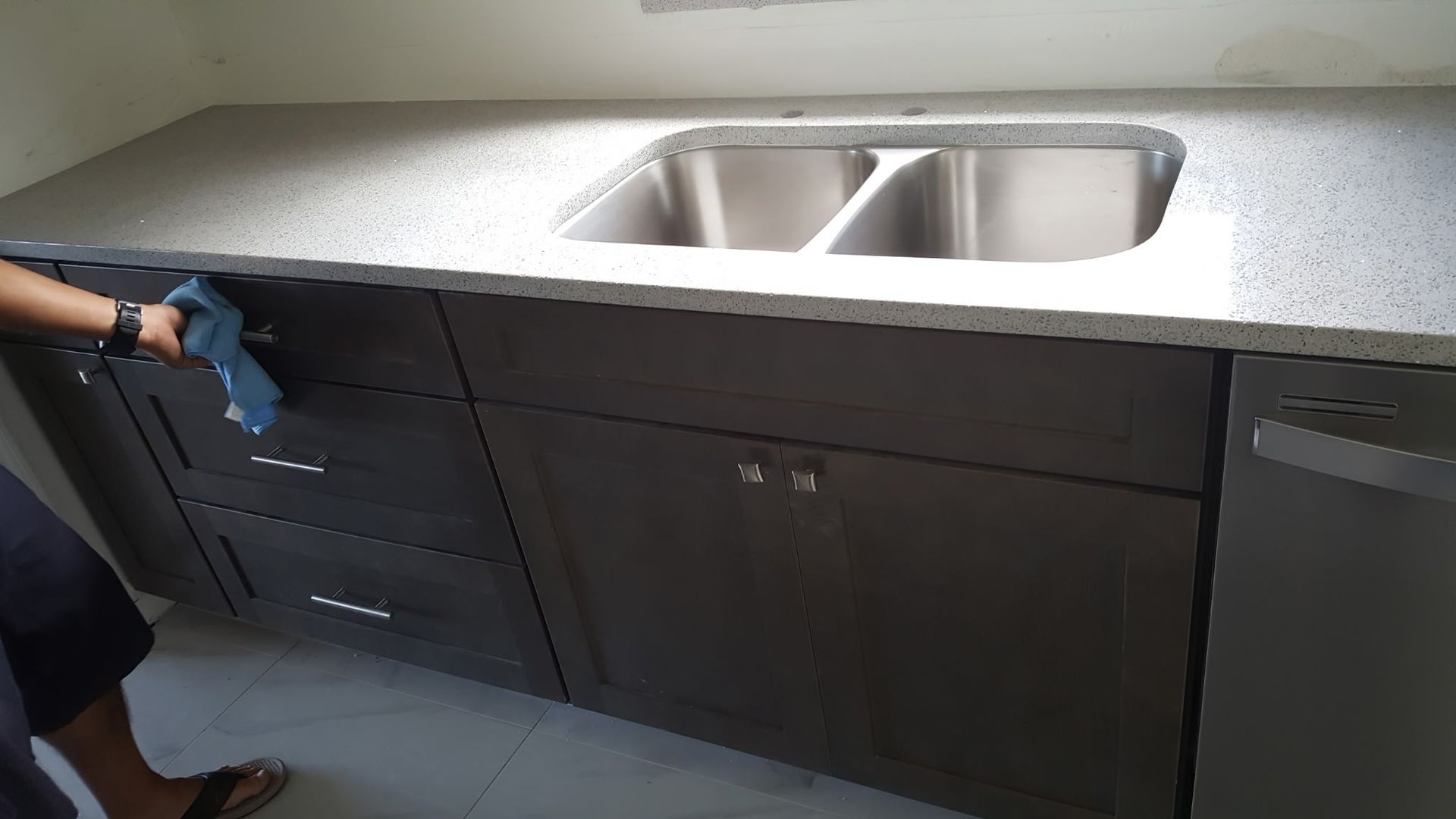 a person wiping a dark brown kitchen cabinet near a stainless steel sink and dishwasher