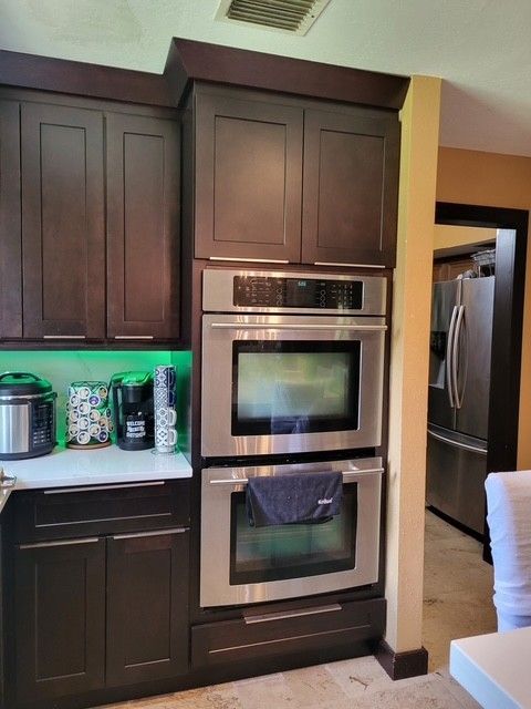 dark brown kitchen cabinets with built-in stainless steel double oven, green under-cabinet lighting, and glimpse of a refrigerator