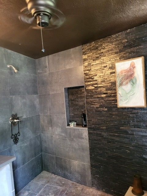 bathroom with gray tiled shower, dark stone accent wall, artwork, and ceiling fan