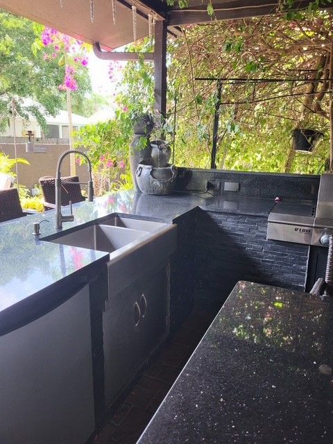 outdoor kitchen with black countertops, sink, grill, and surrounding foliage