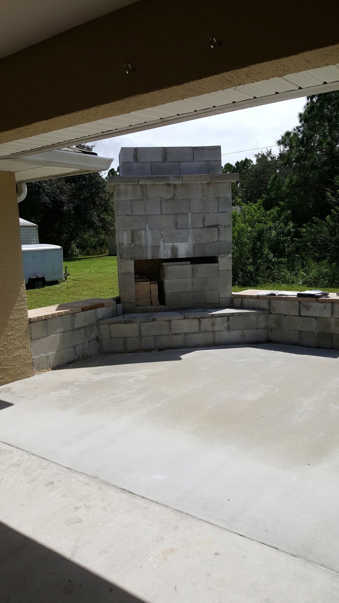outdoor fireplace under a patio roof; built from grey cinder blocks on a concrete patio, green trees in background
