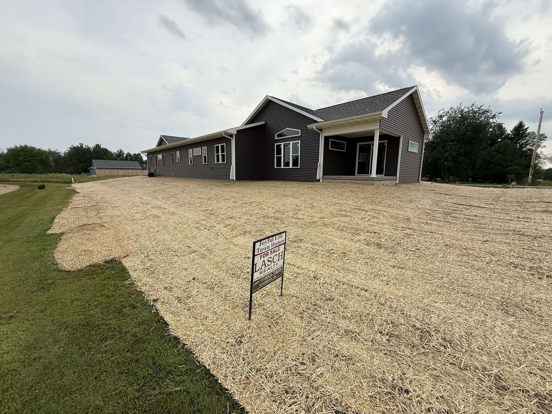 A dark grey, single-story house with a white-trimmed porch stands on a large, gravel-covered lot under a cloudy sky.