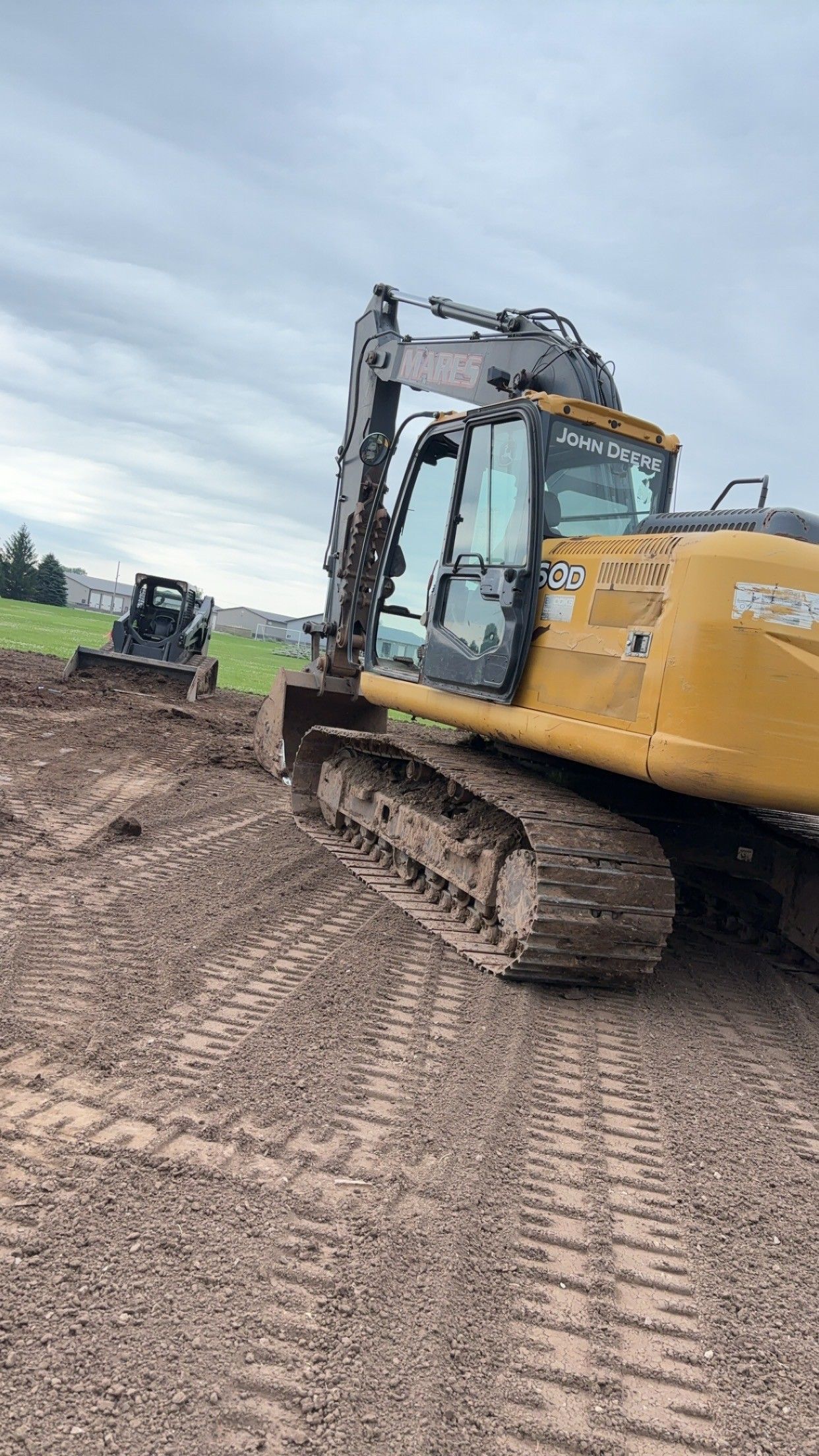 A yellow tracked excavator sits on a dirt construction site with a small skid steer loader visible in the background.