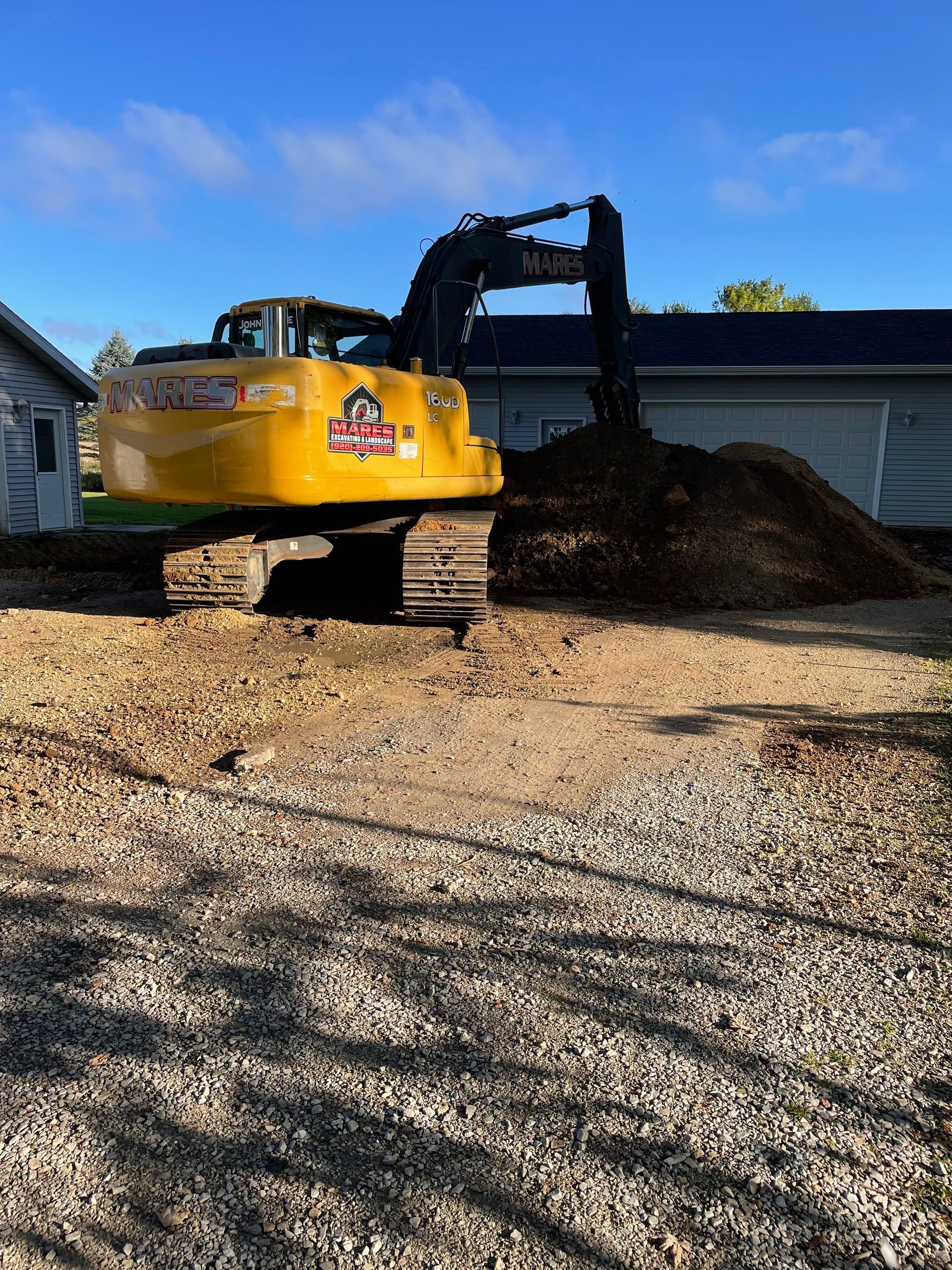 A yellow excavator is working on a gravel road in front of a house.
