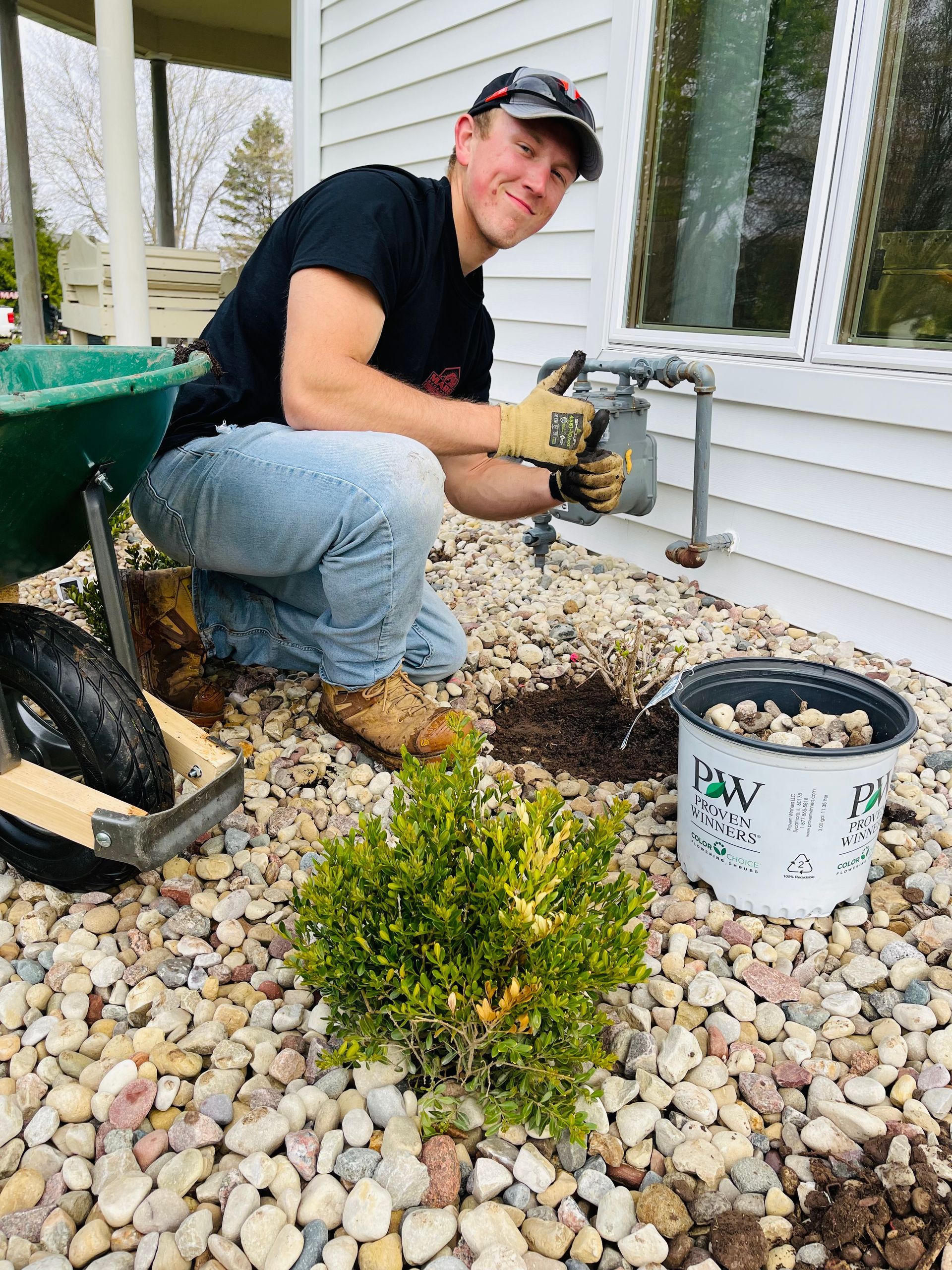A man is kneeling down in front of a house with a wheelbarrow.